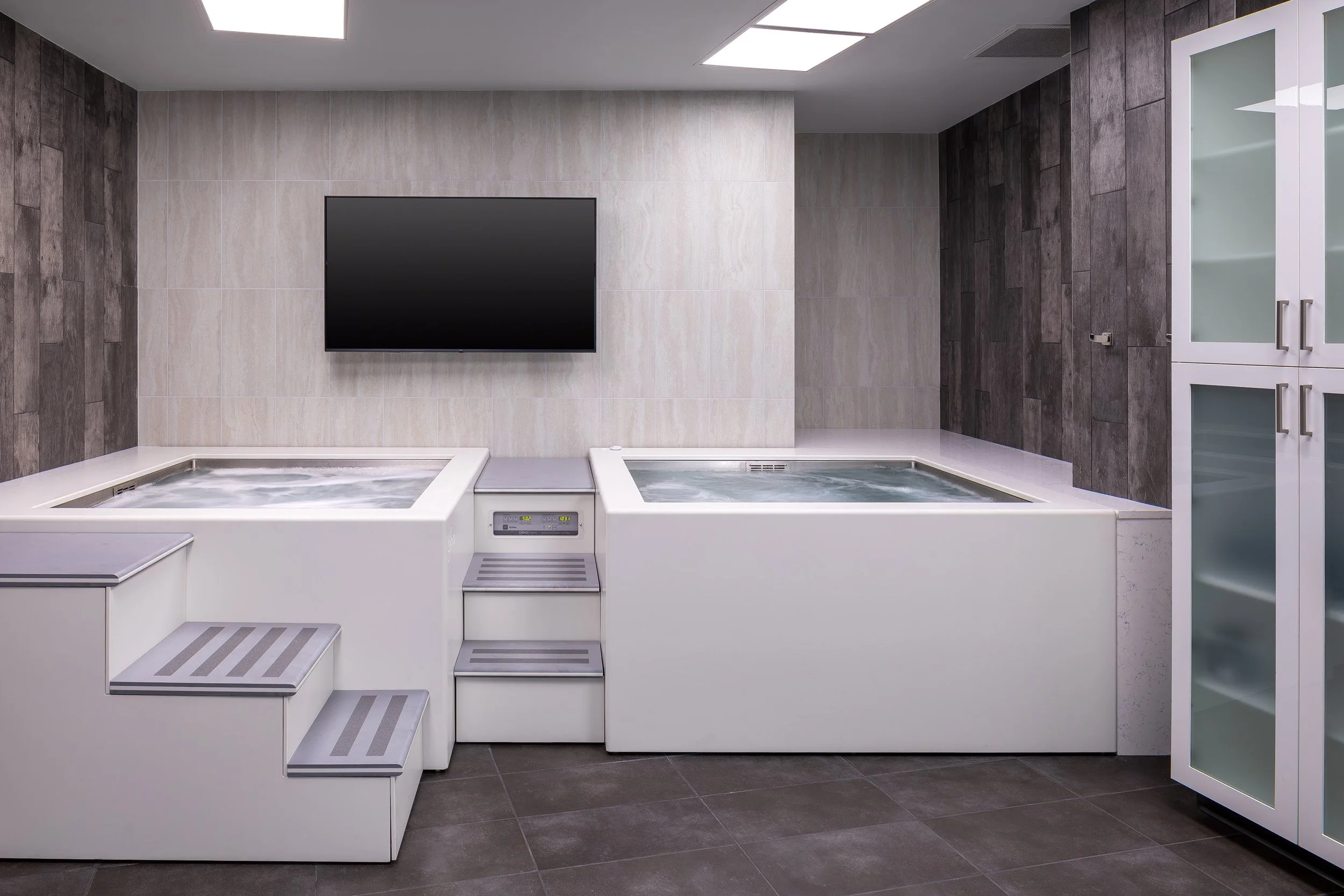 A modern room with two white hot tubs side by side, a set of steps leading up to them, a flat-screen TV on the wall, and glass-front cabinets on the right. The space has tiled floors and walls.