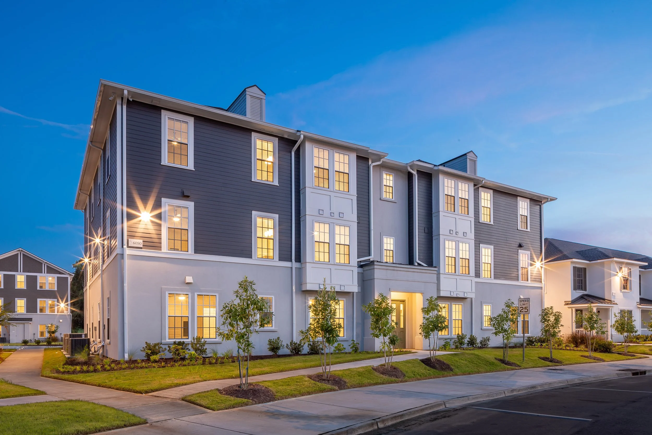 Modern three-story apartment building with large windows, gray and white exterior, well-maintained landscaping, and streetlights illuminating the area at dusk.