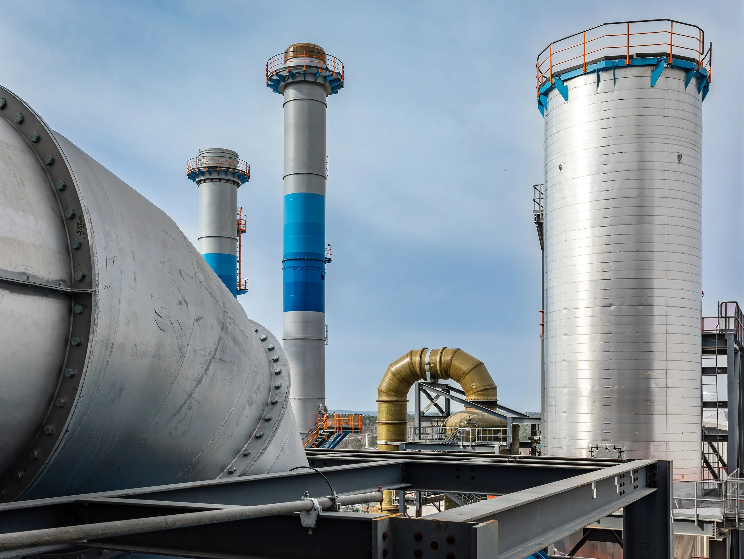 A large industrial metal structure with pipes, with a blue clouded sky behind.
