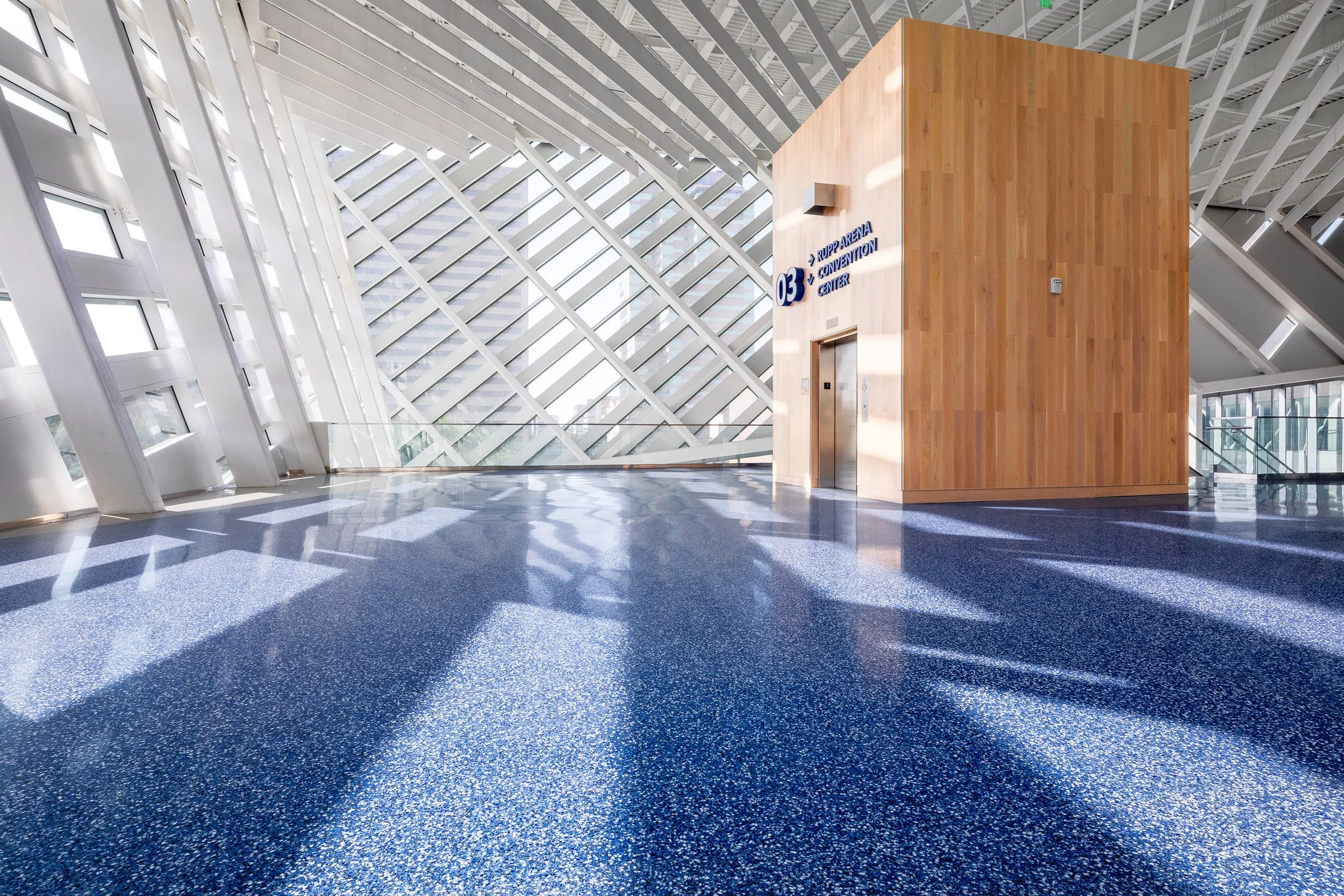 Large, bright lobby with blue speckled floors and wood-paneled elevator showcases Rupp Arena modern architecture and angled glass windows for natural light.
