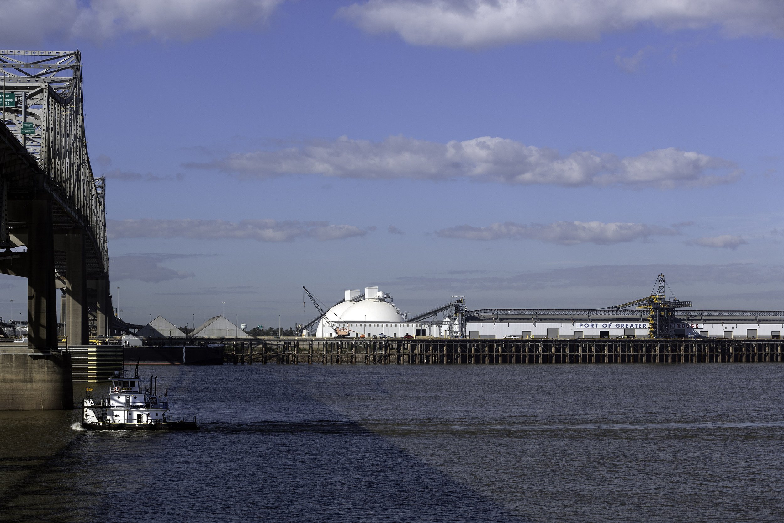 A tugboat on a river near a large bridge and an industrial facility with white domes in the background.