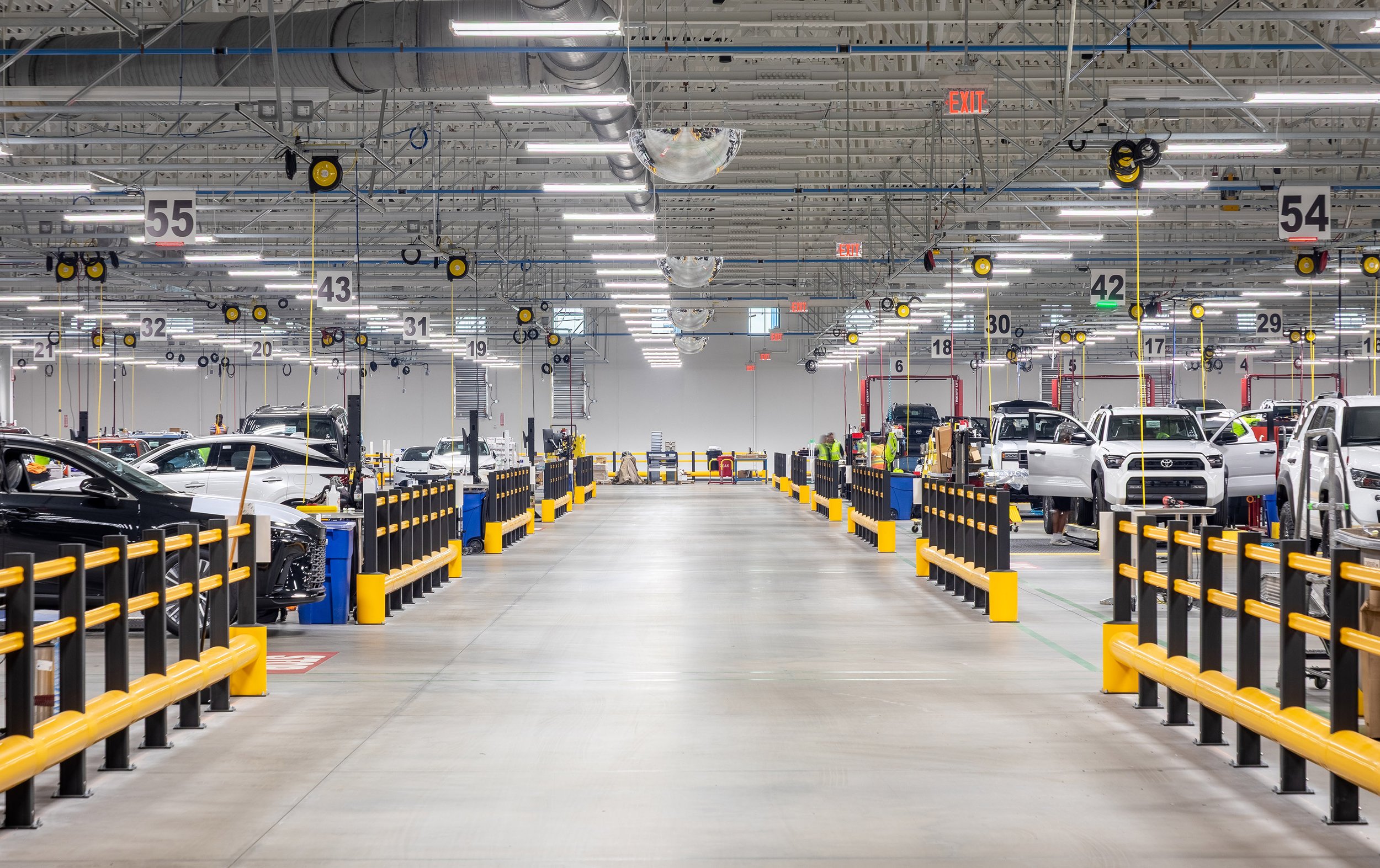 Wide indoor view of an automotive service center or factory, with cars parked in rows on each side, technicians working, bright lighting, numbered signs overhead, and yellow safety barriers lining the work areas.