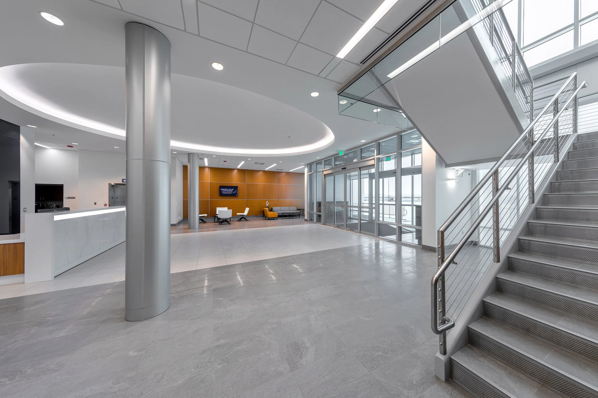 A modern architectural office lobby with gray tile floors, a staircase on the right, glass walls, a reception desk on the left, and a lounge area with chairs and a TV in the background.