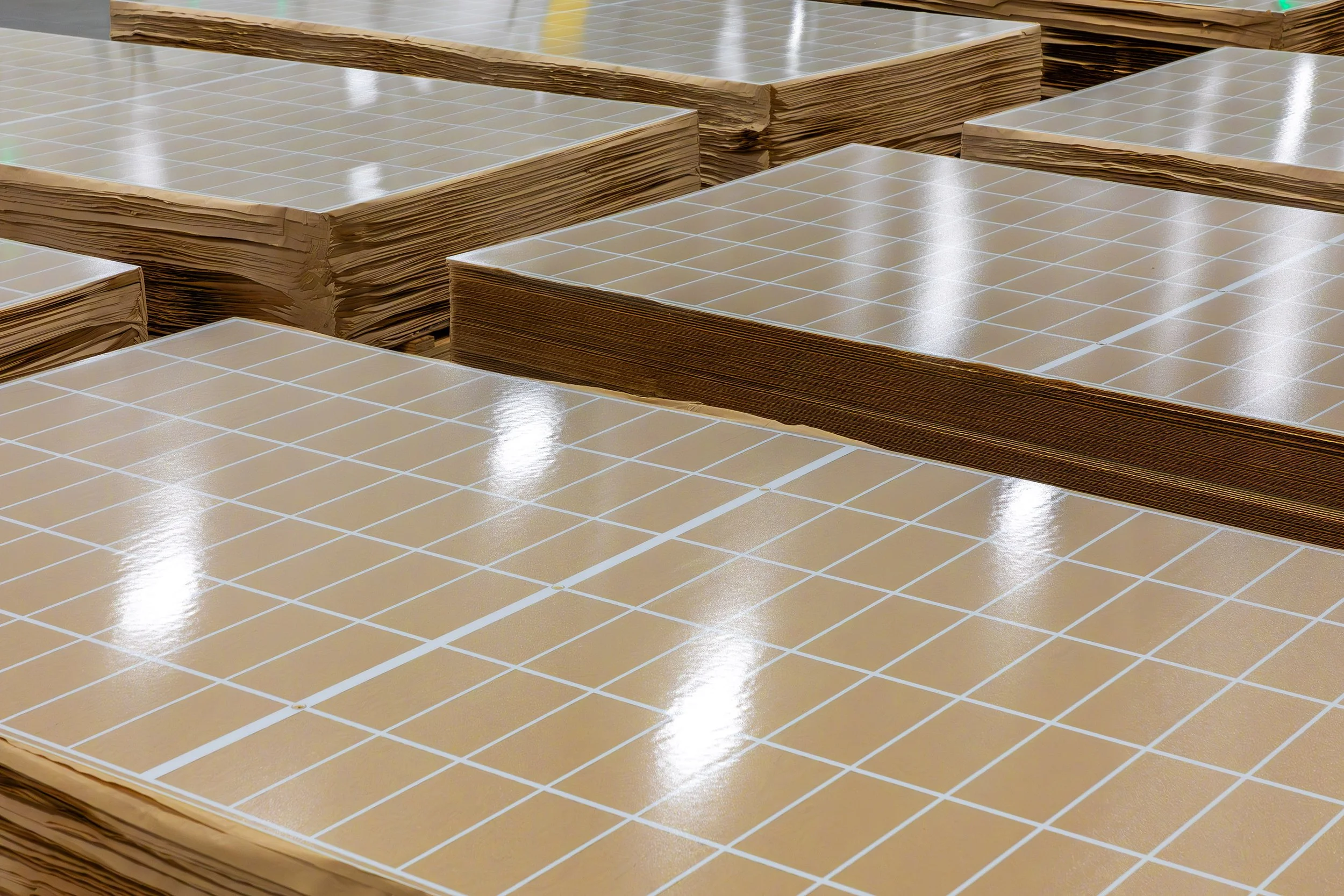 Stacks of large, glossy sheets with a grid pattern, likely for solar panels, are arranged in rows inside an industrial warehouse.