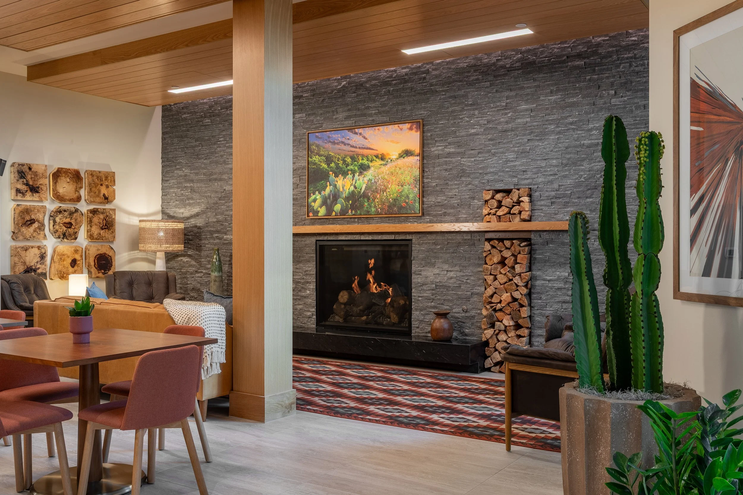 A cozy modern living space with a fireplace, stacked firewood, a cactus plant, wooden dining table with pink chairs, and wall art, featuring warm lighting and a patterned rug.