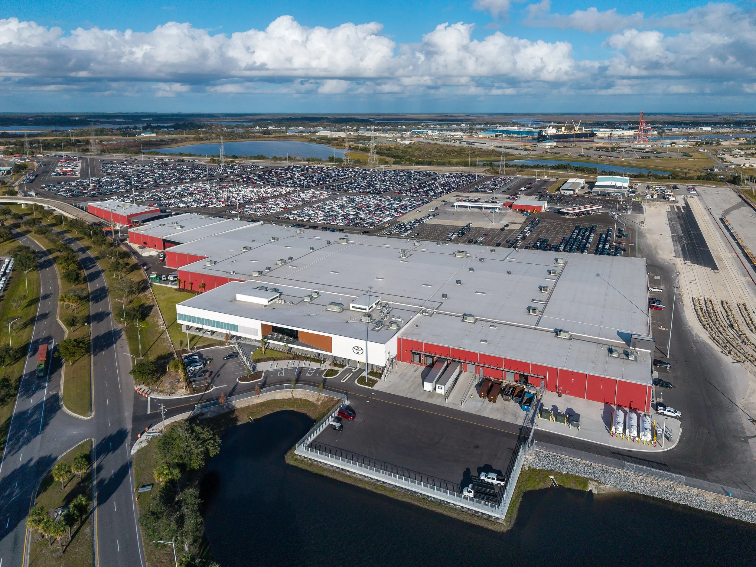 Aerial view of a large industrial facility with a white and red roof, surrounded by parking lots, cars, a body of water, and roads, under a partly cloudy sky.