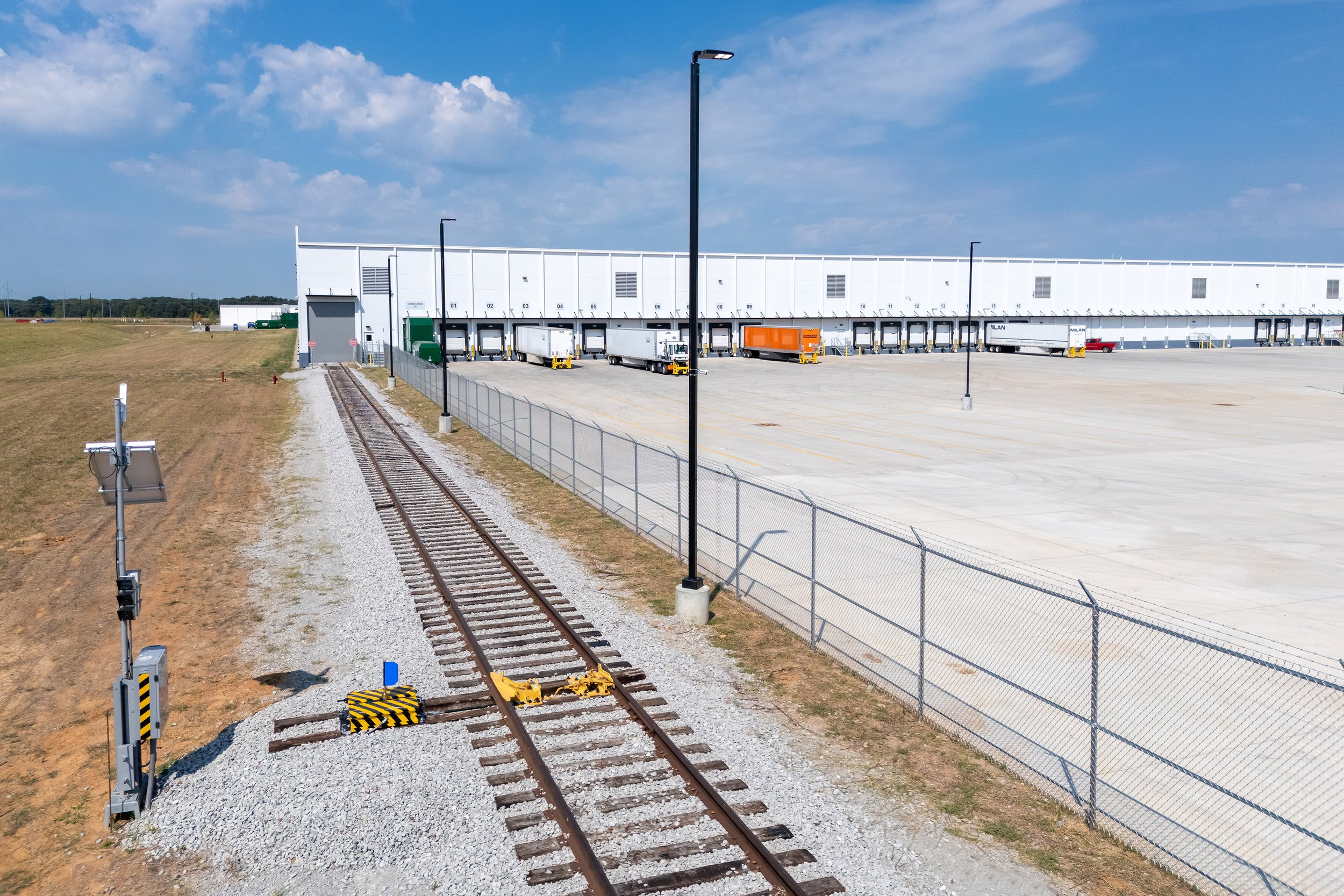 A railway track ends at an industrial warehouse with loading docks and trucks parked under a blue sky.