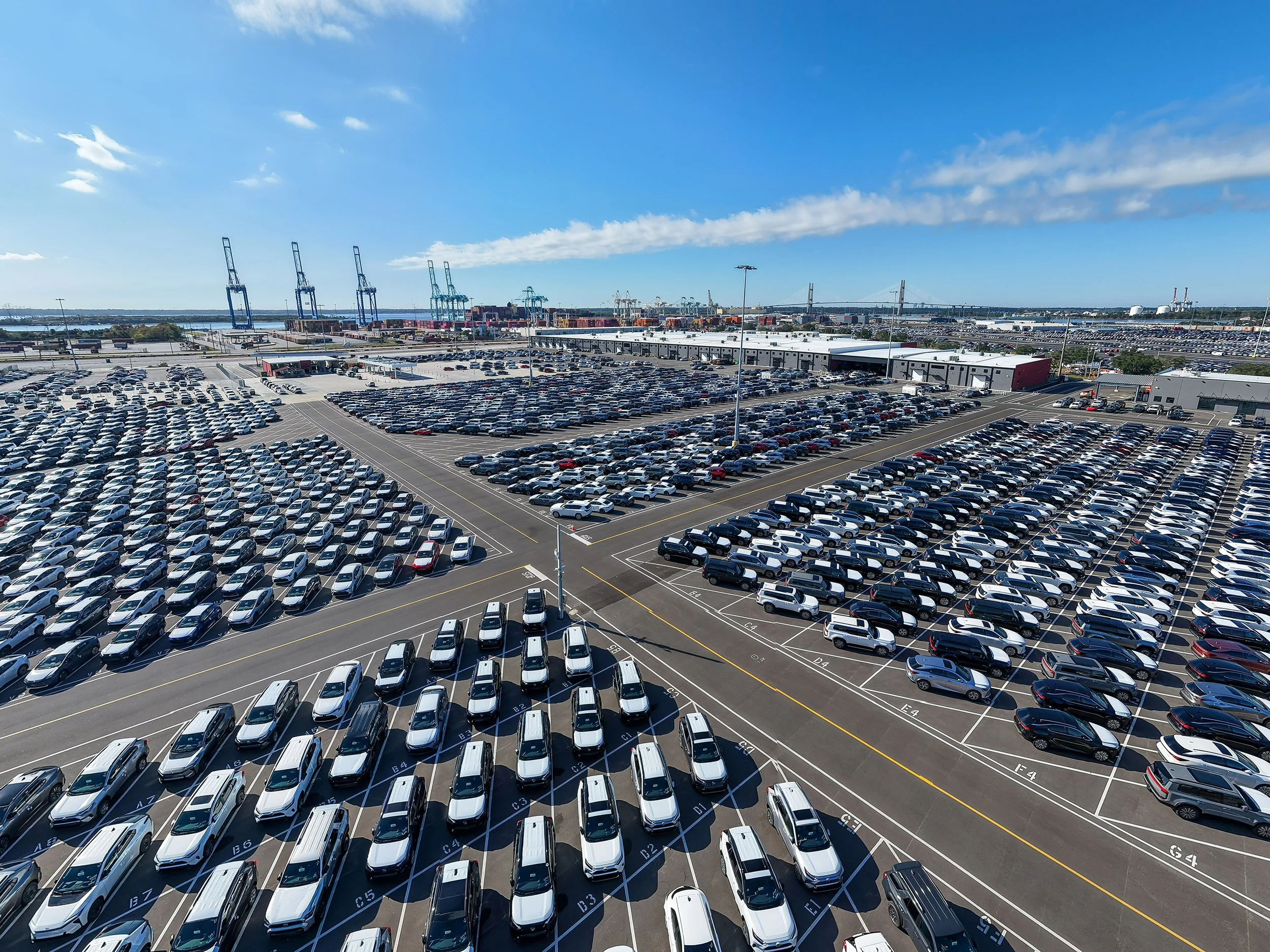 Rows of parked cars fill a large lot near shipping cranes at a port under a clear blue sky, suggesting a car import or export facility with hundreds of vehicles organized in neat lines.