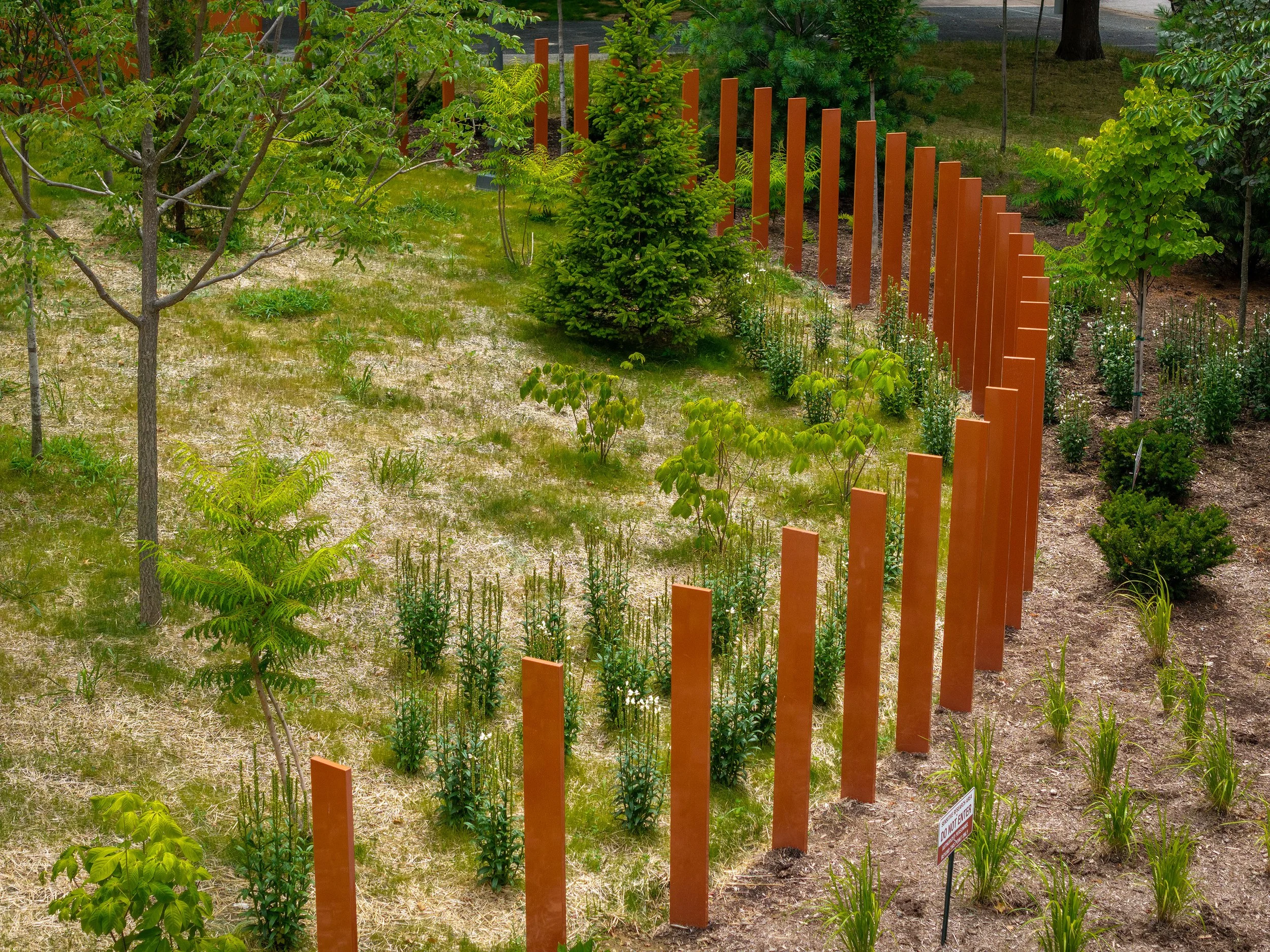 A curved row of tall, rust-colored metal posts stands in a grassy garden with trees, shrubs, and mulch, blending landscape and architecture to create an artistic outdoor installation surrounded by greenery.