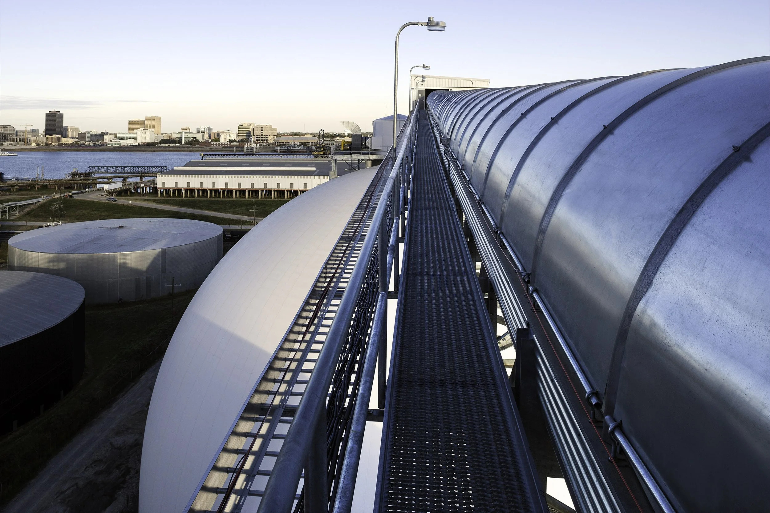 Industrial walkway with metal railings and structures, city skyline and water in the background under blue sky.