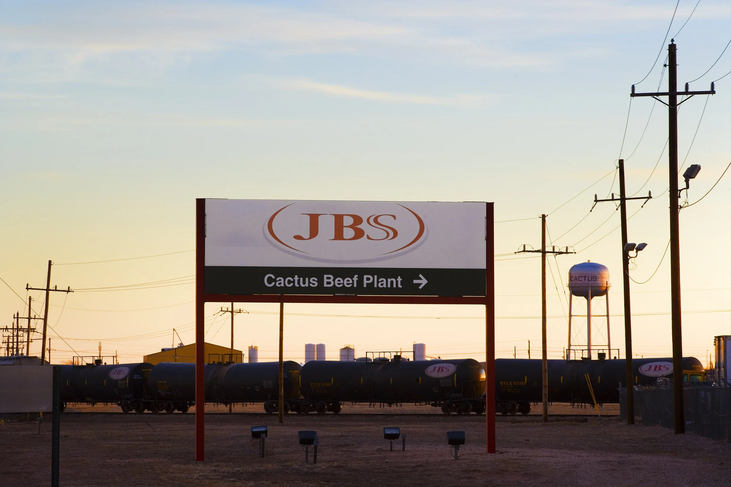 Sign for JBS Cactus Beef Plant with tanker cars and power lines at sunset.