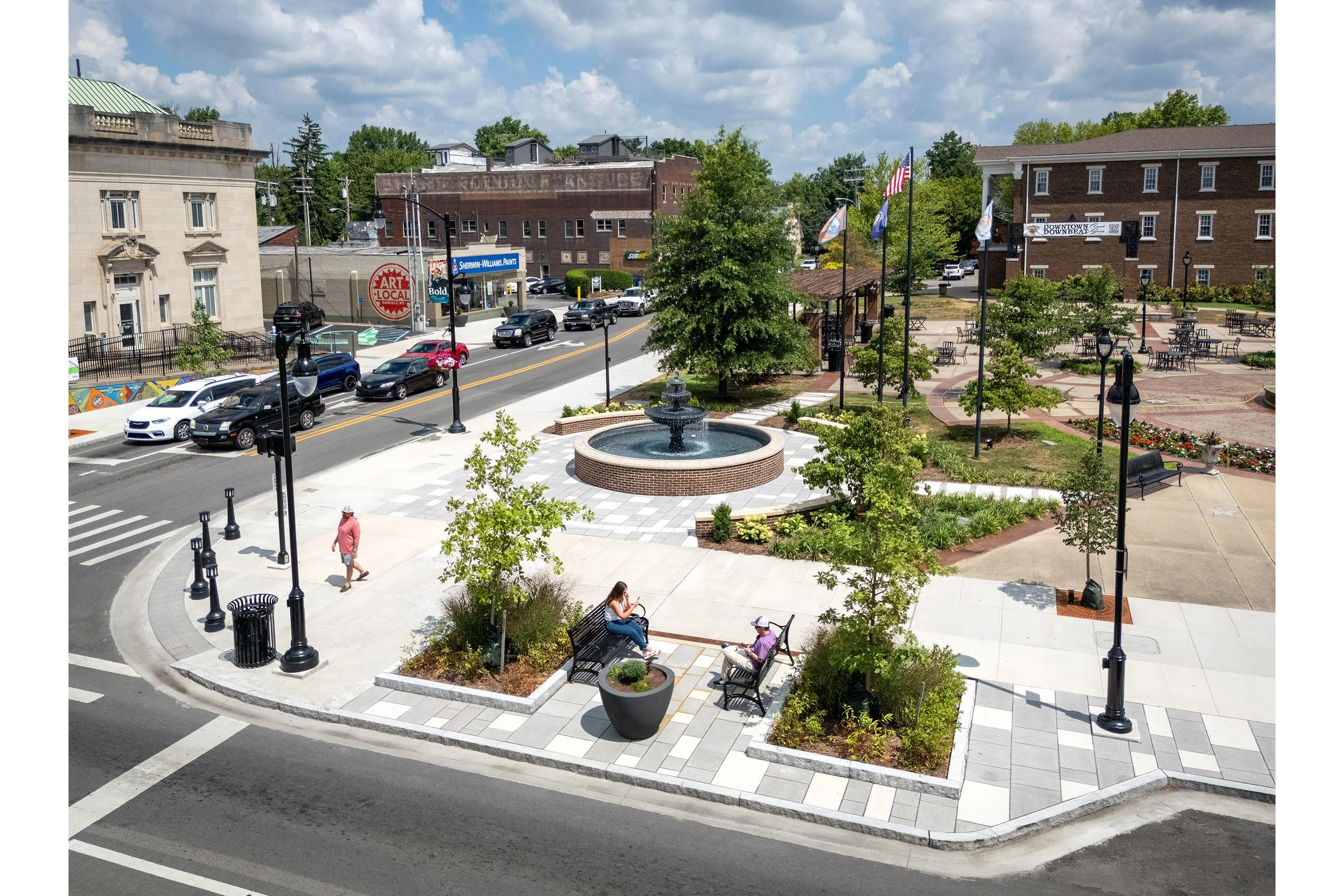 A small urban park with a central fountain, benches, trees, and people walking adds charm to the historic streetscape on a sunny day.
