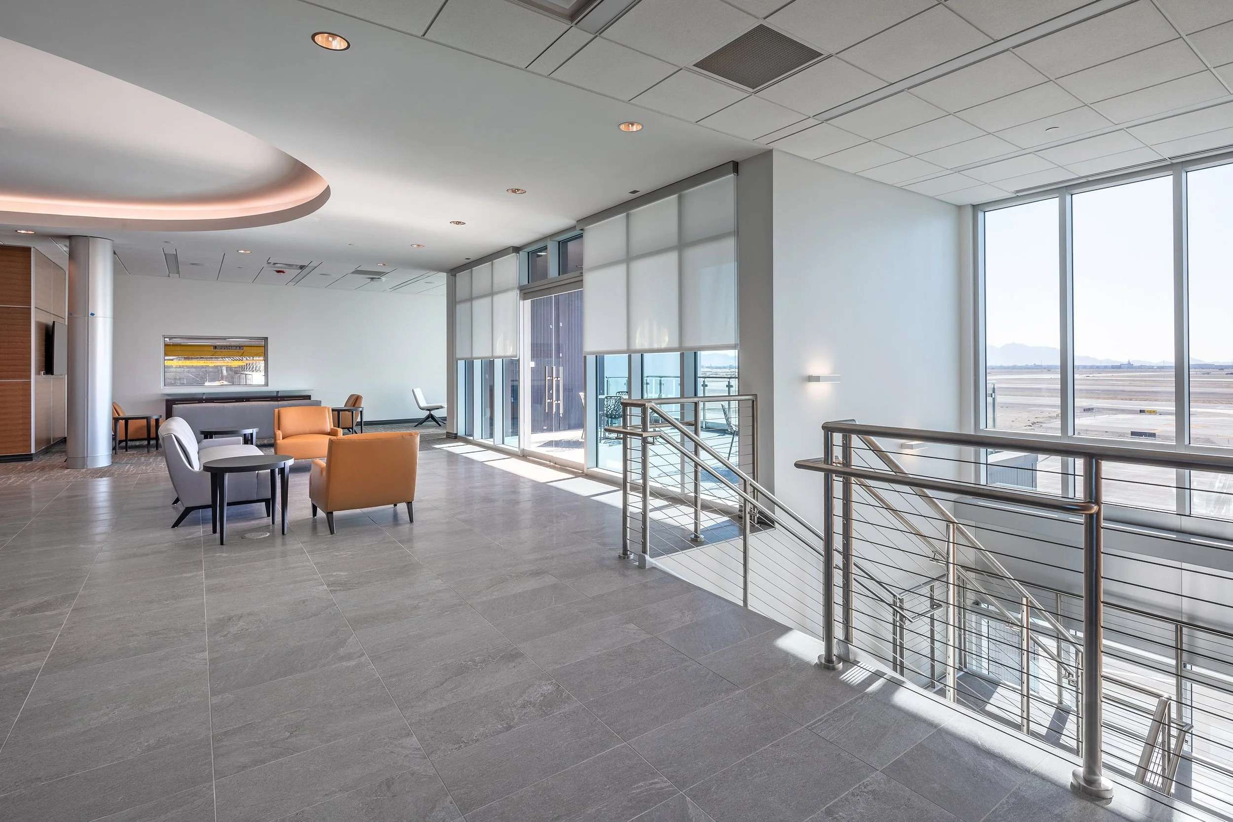 Modern airport lounge with large windows and grey tile floor, captured in stunning architectural photography. Orange and grey chairs fill the bright, spacious area as sunlight streams in, revealing Arizona runways just beyond the expansive glass.