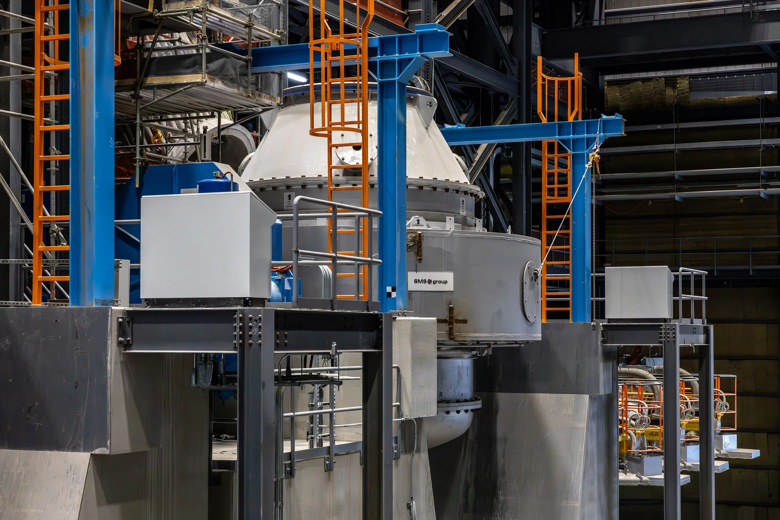 Large scale industrial process with gray and blue metal components processes metals, surrounded by orange safety railings and ladders, inside a spacious manufacturing facility.