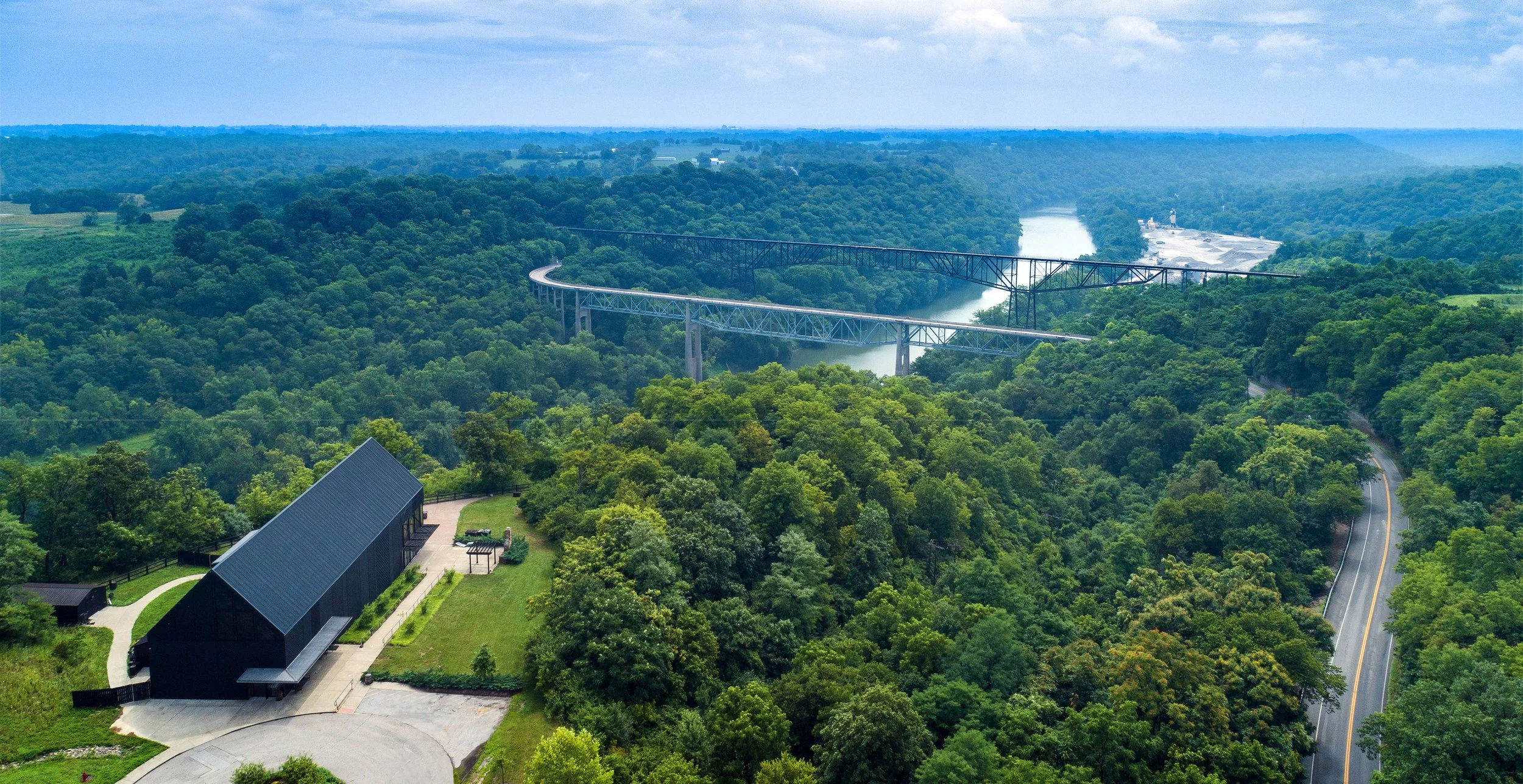 A scenic landscape with a winding river, surrounded by lush green forests, a large black building in the foreground, and a long bridge crossing the river in the background under a partly cloudy sky.