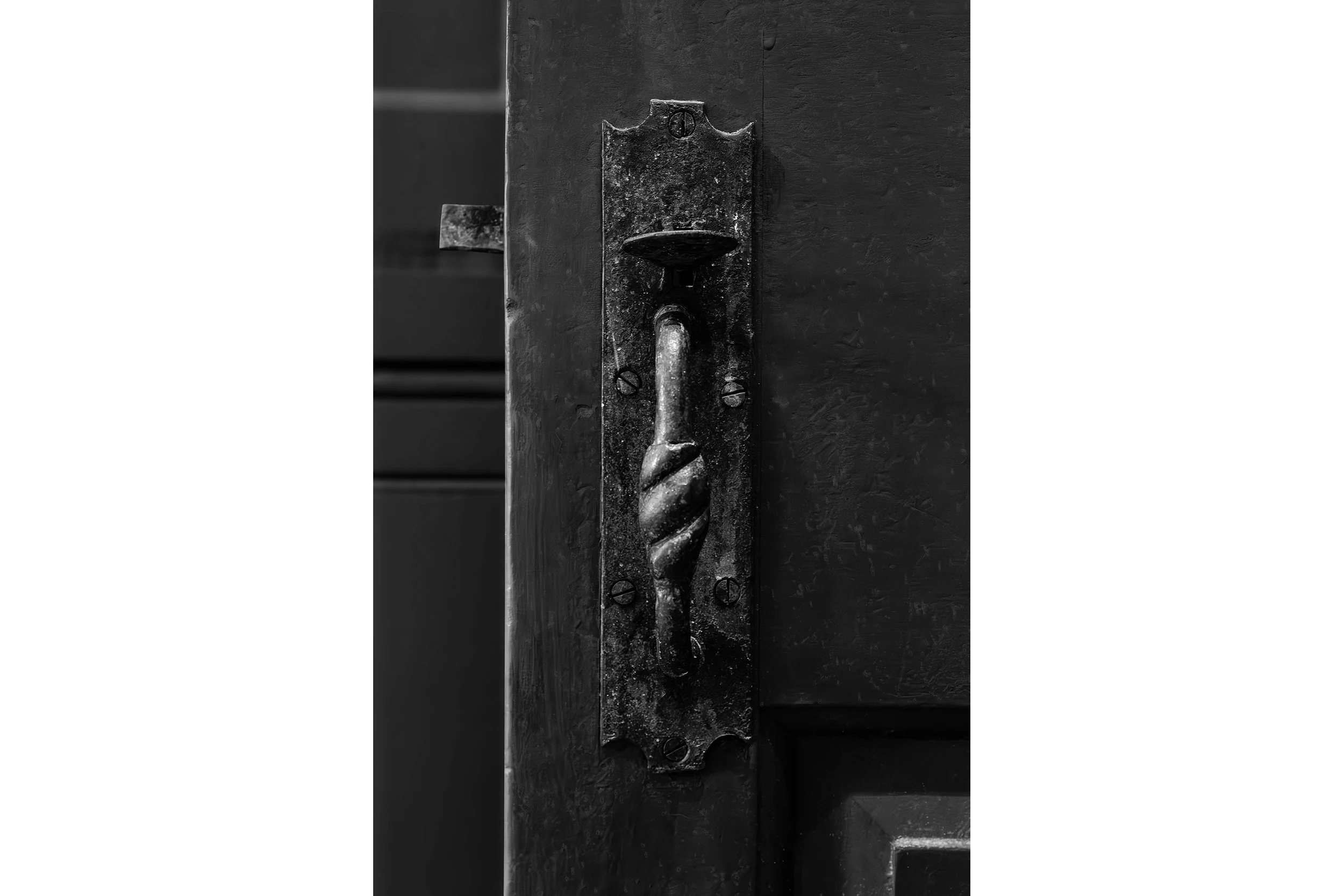 Black and white photo of an old, ornate metal door handle on a dark wooden door of a historic Shaker building.