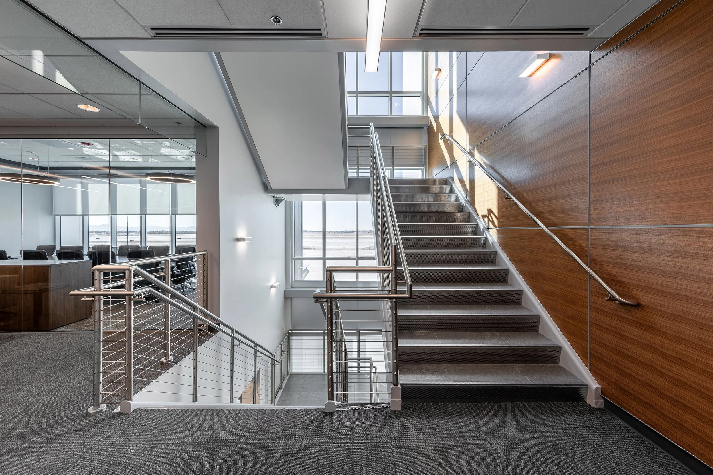 A glass door opens to an Arizona balcony with metal chairs and tables, overlooking a vast, sunny landscape and distant mountains beneath a clear blue sky—perfect for architectural photography as shadows dance across the floor.