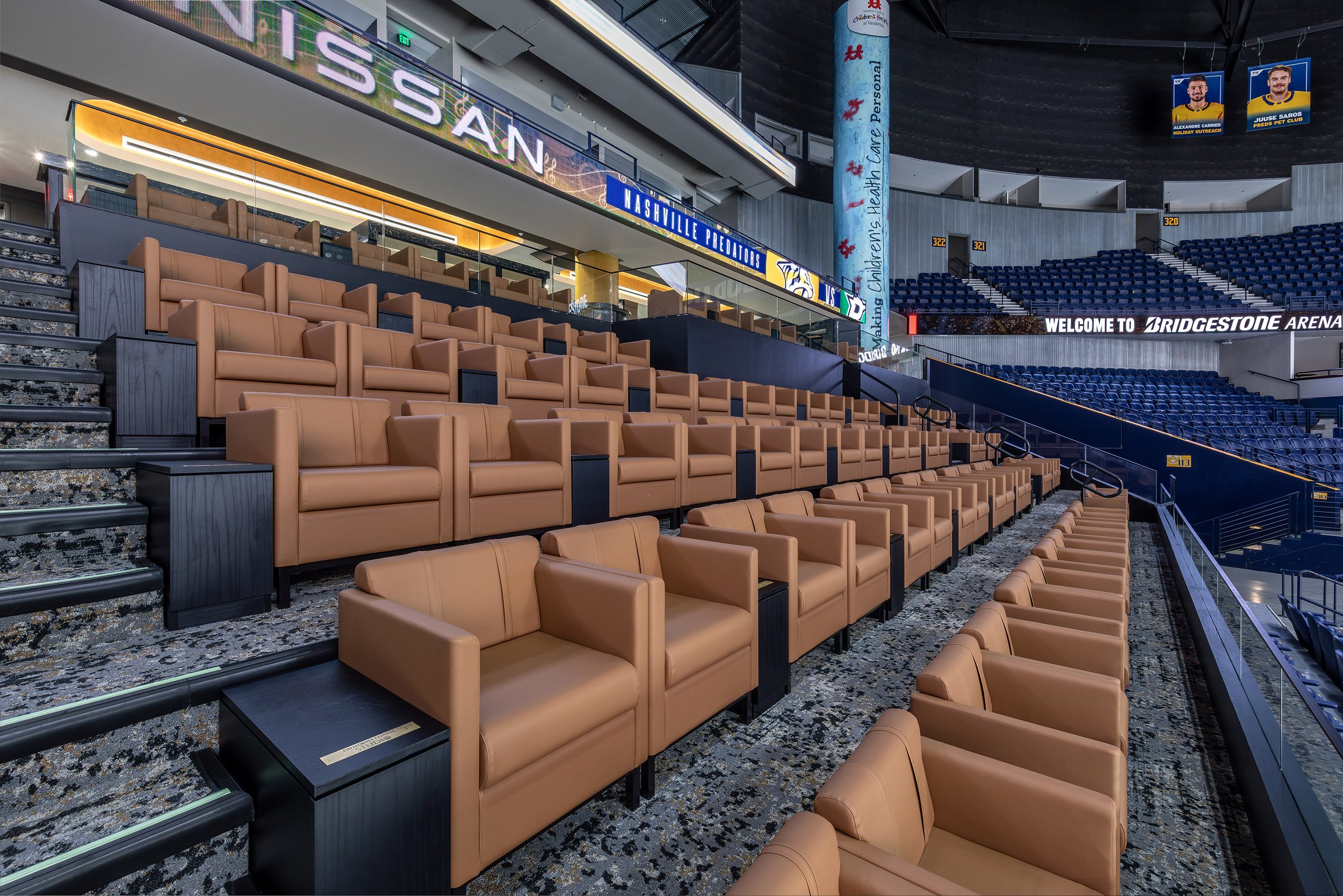 Rows of plush tan leather armchairs are arranged in a luxury club seating section inside a large indoor sports arena, with blue stadium seats and advertising signs visible in the background.