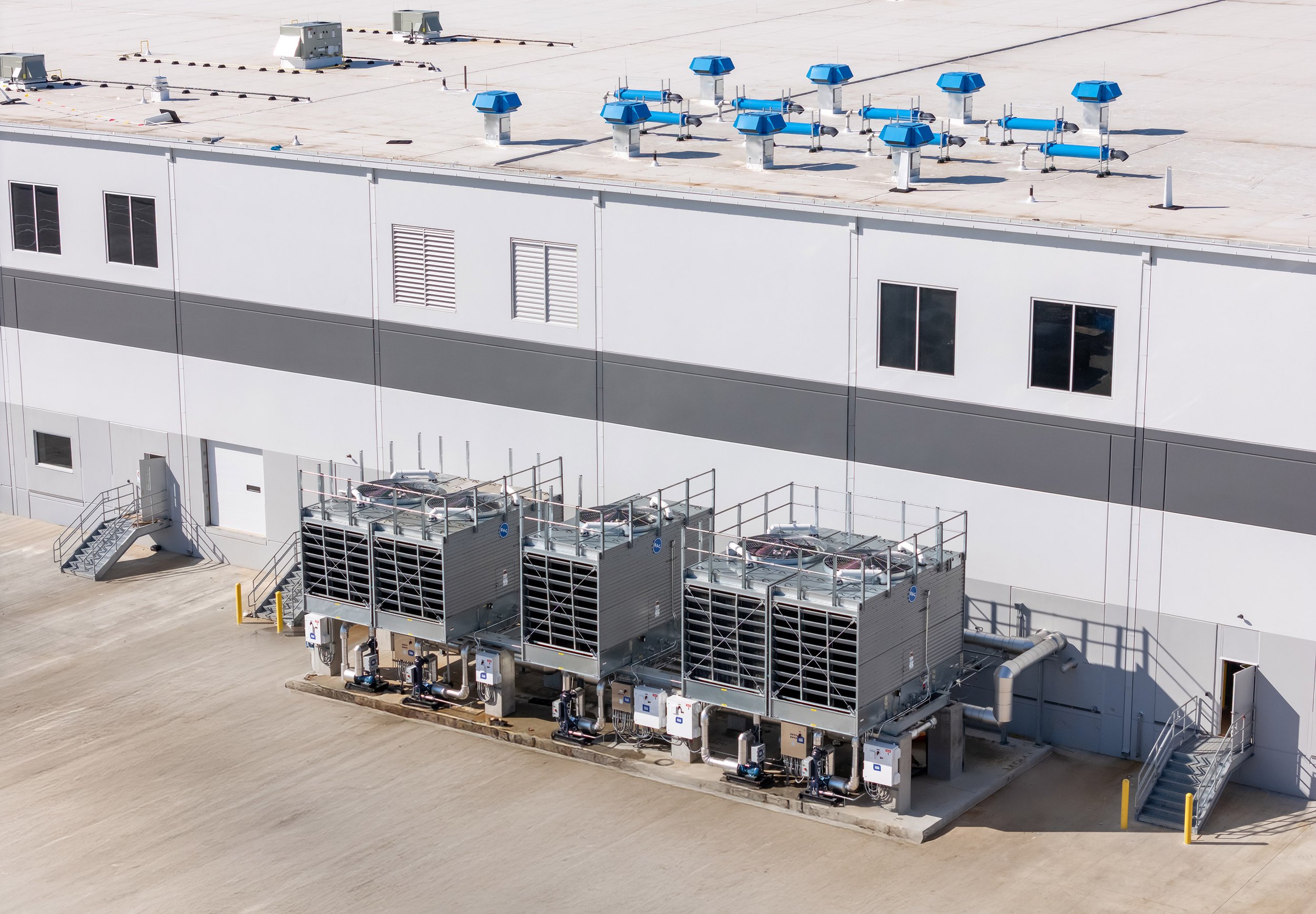 Three large industrial air conditioning units are installed outside a commercial building in Dallas, Texas. The flat roof features multiple blue ventilation systems, with several windows and exterior stairs on the building’s facade.