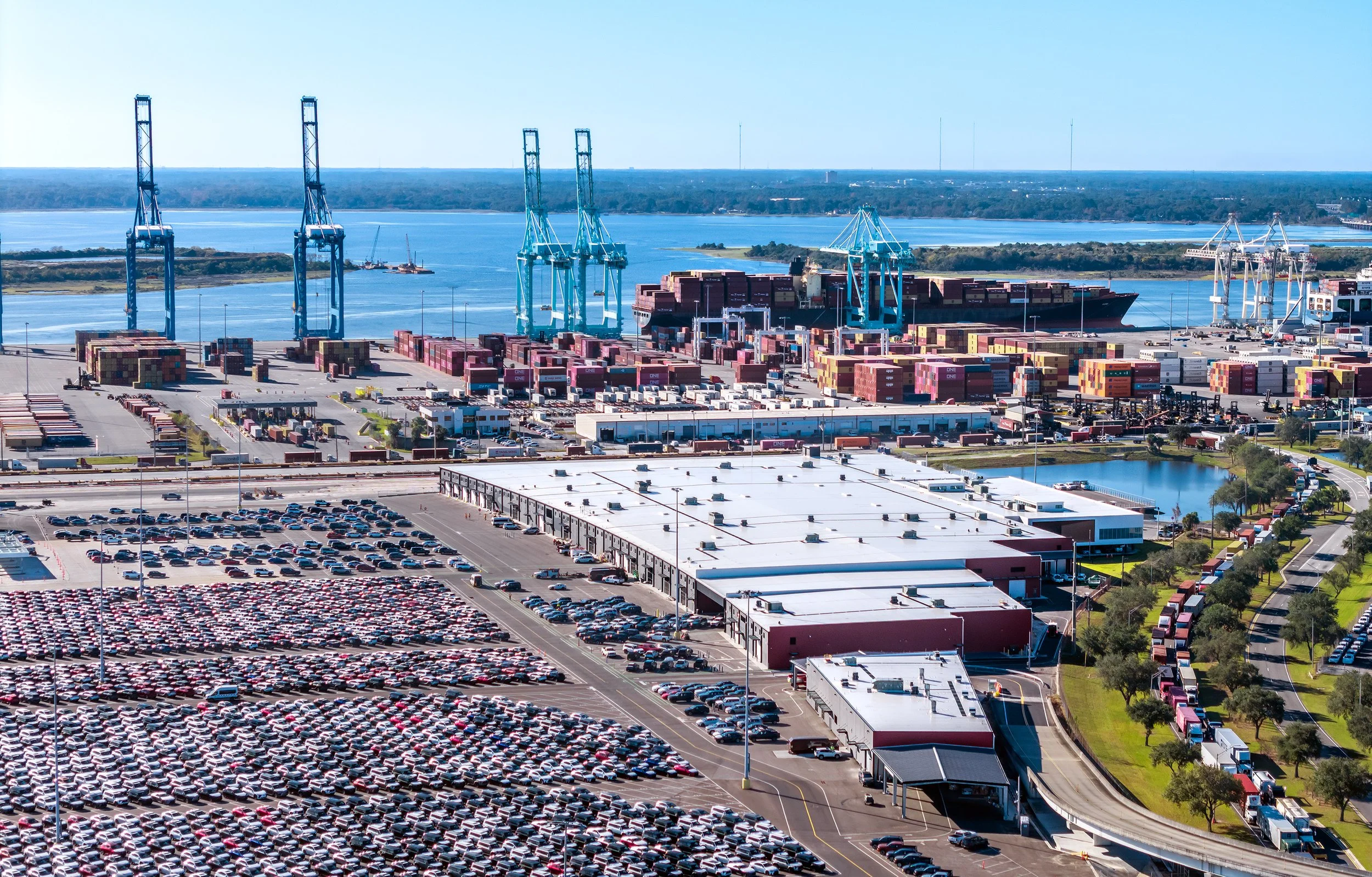 Aerial view of a large port terminal with cargo ships, cranes, and stacked shipping containers near a body of water. There are parking lots filled with cars and a road with trucks on the right side.