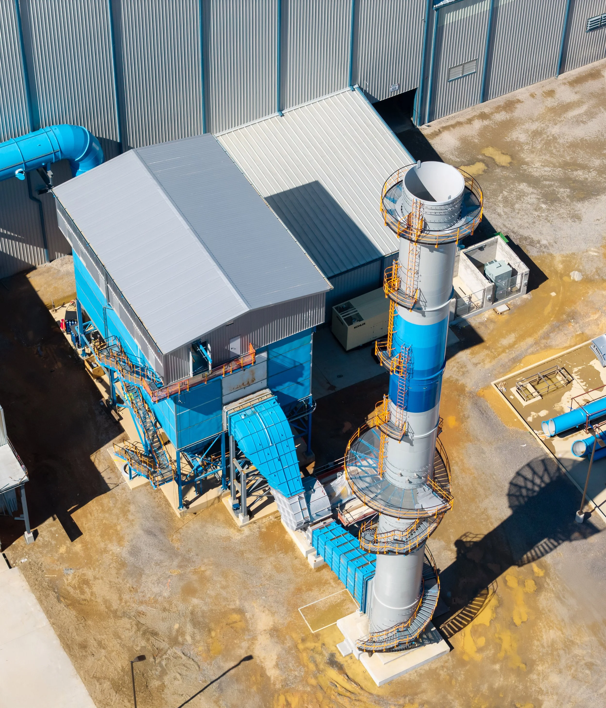 Aerial view of an industrial facility showcasing architectural details—a large silver and blue building with connected pipes, a tall chimney encircled by metal staircases, and railings. The paved area is bordered by a corrugated metal fence.