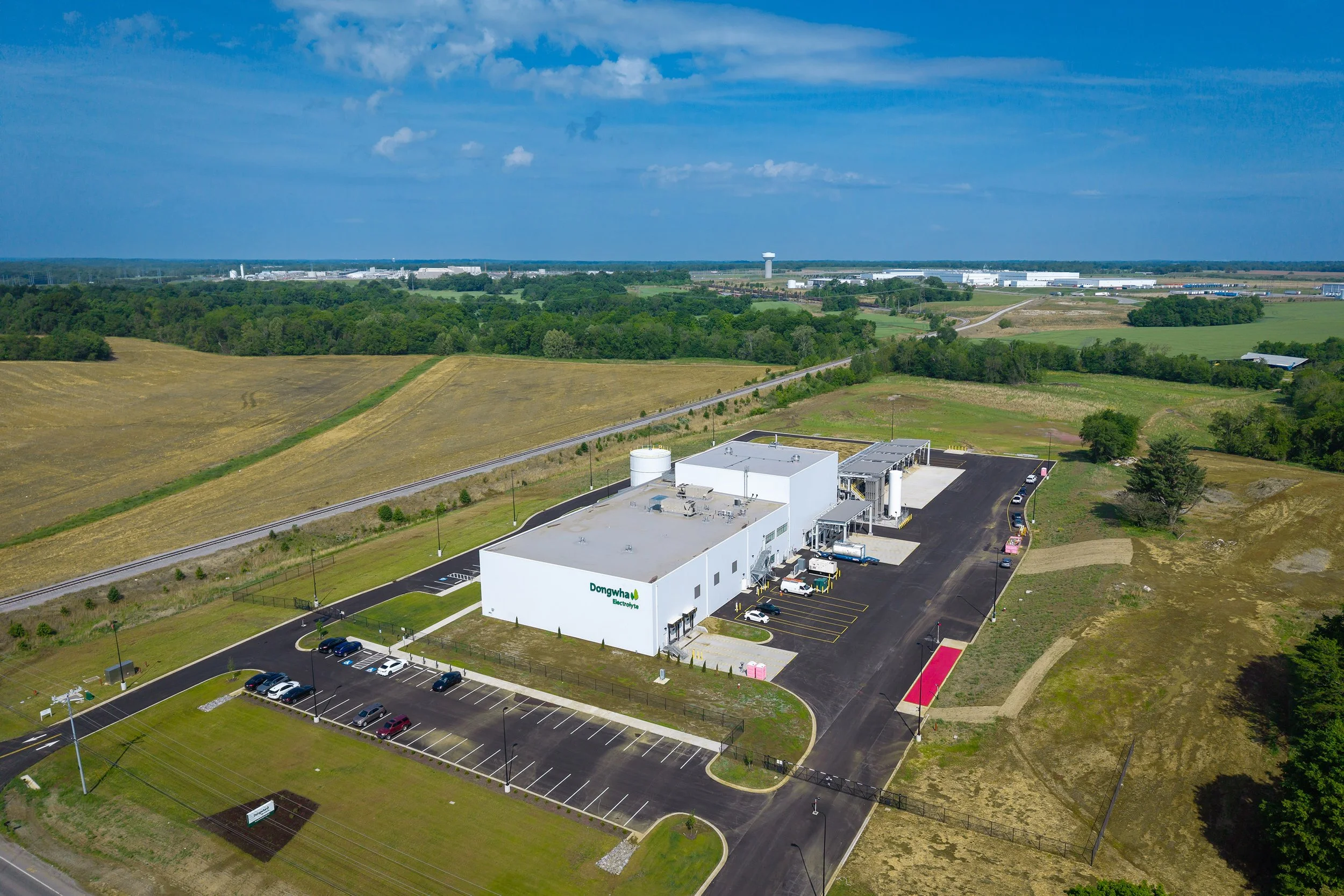 Aerial view of a large white industrial building with Domtar signage, showcasing an architectural design surrounded by fields and trees, a parking lot with cars, and a road running alongside the property under a blue sky.