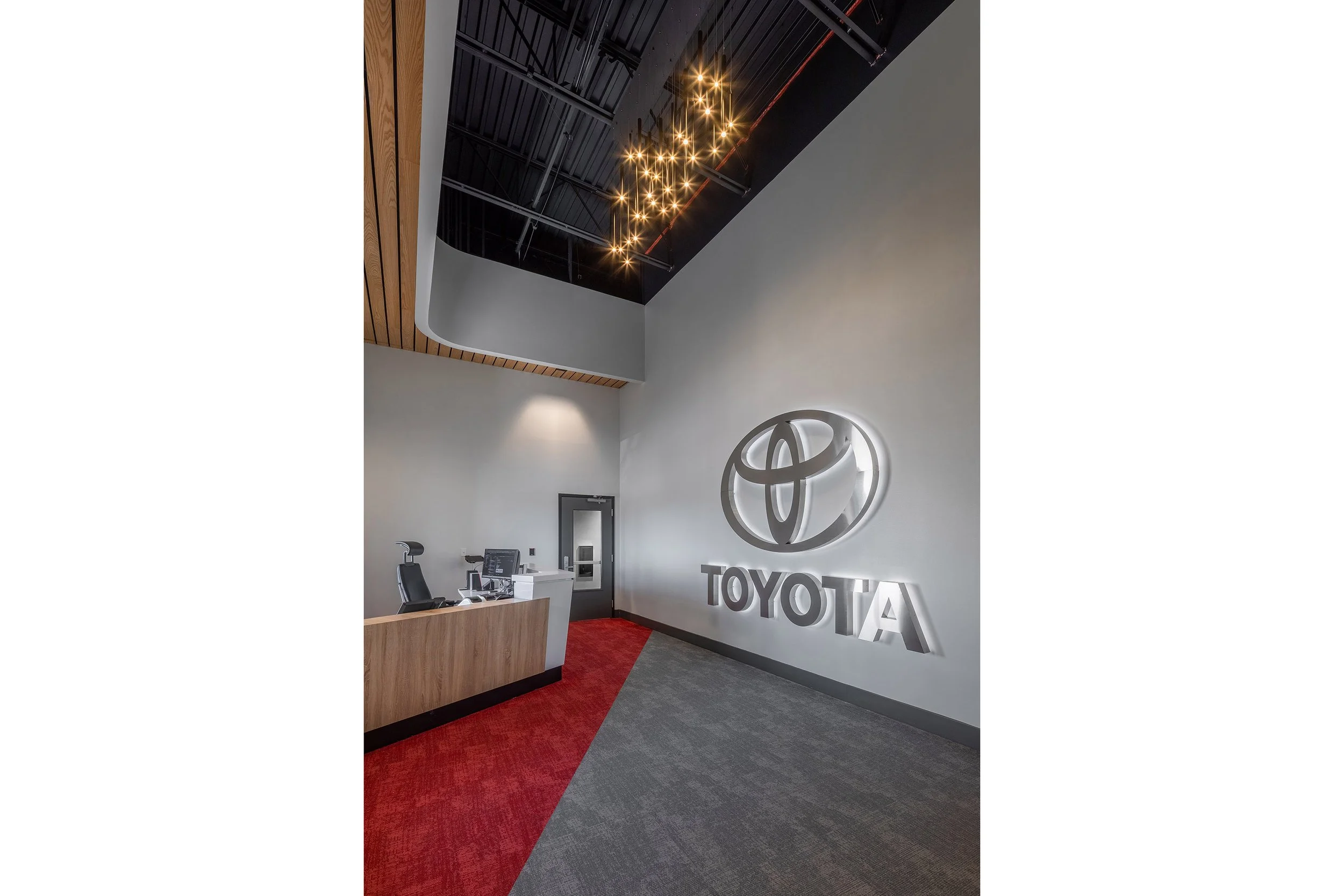 A modern office lobby with a reception desk, red and gray carpet, a wall-mounted illuminated Toyota logo, and a cluster of pendant lights hanging from the high ceiling.