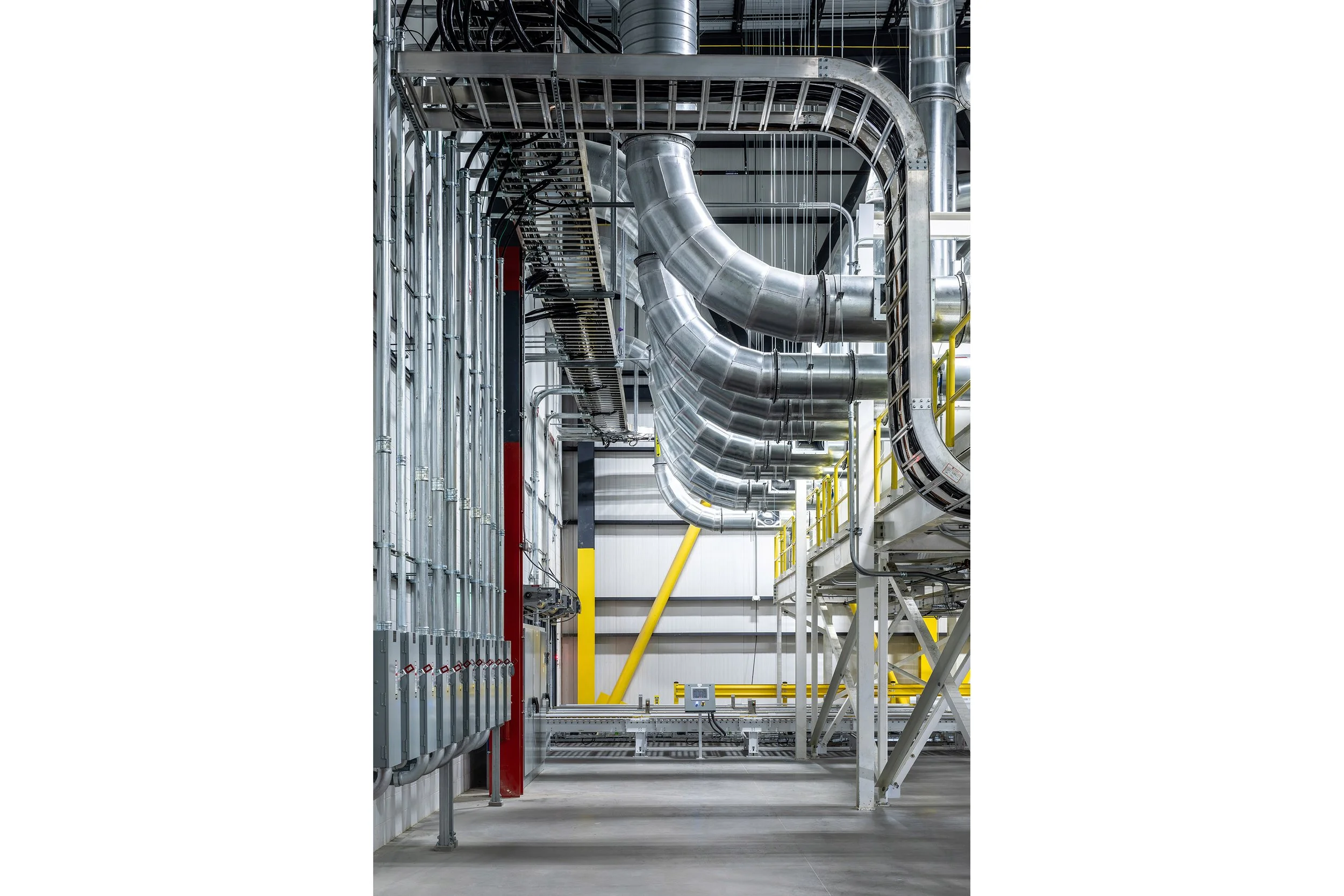 Metal pipes and ducts run along the ceiling and walls of this industrial facility with polished concrete floors and yellow structural beams.