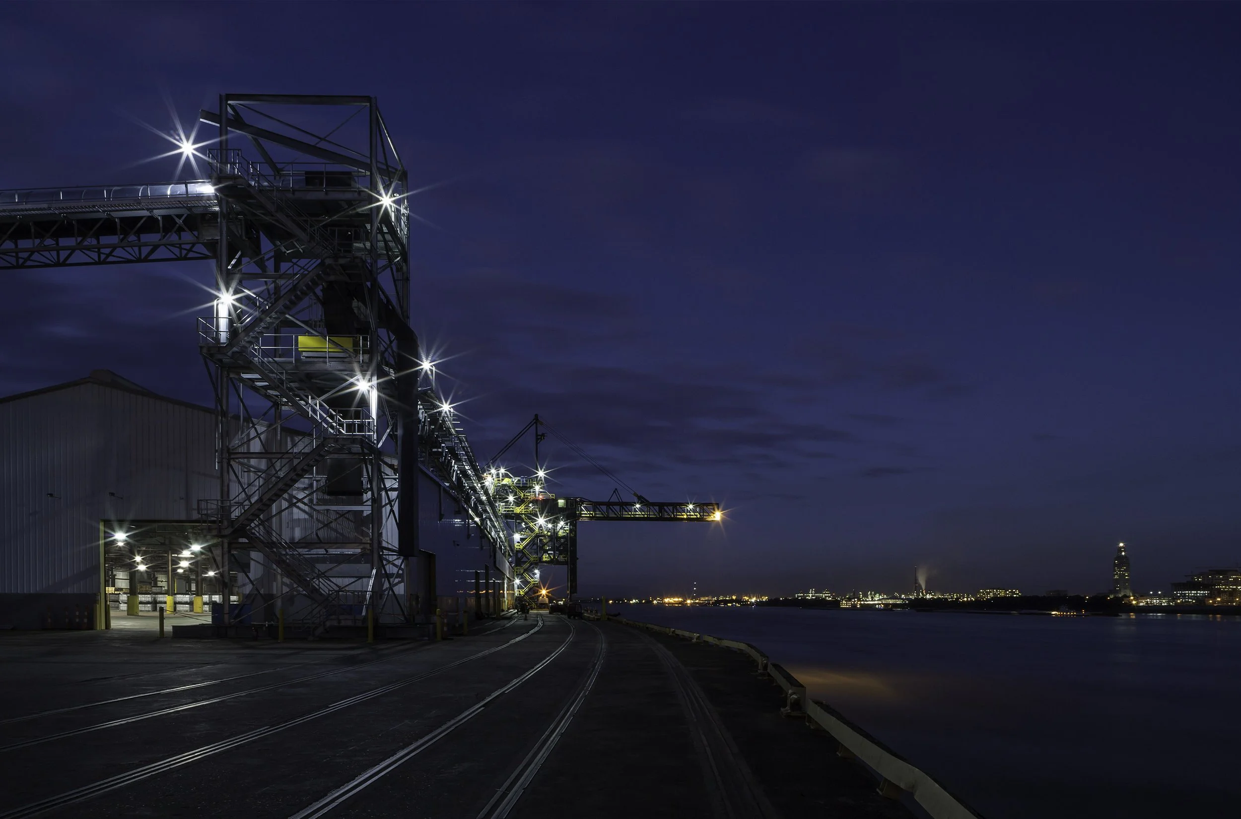 Industrial port facility with conveyor belts and lights at night, next to river with city lights in the distance.