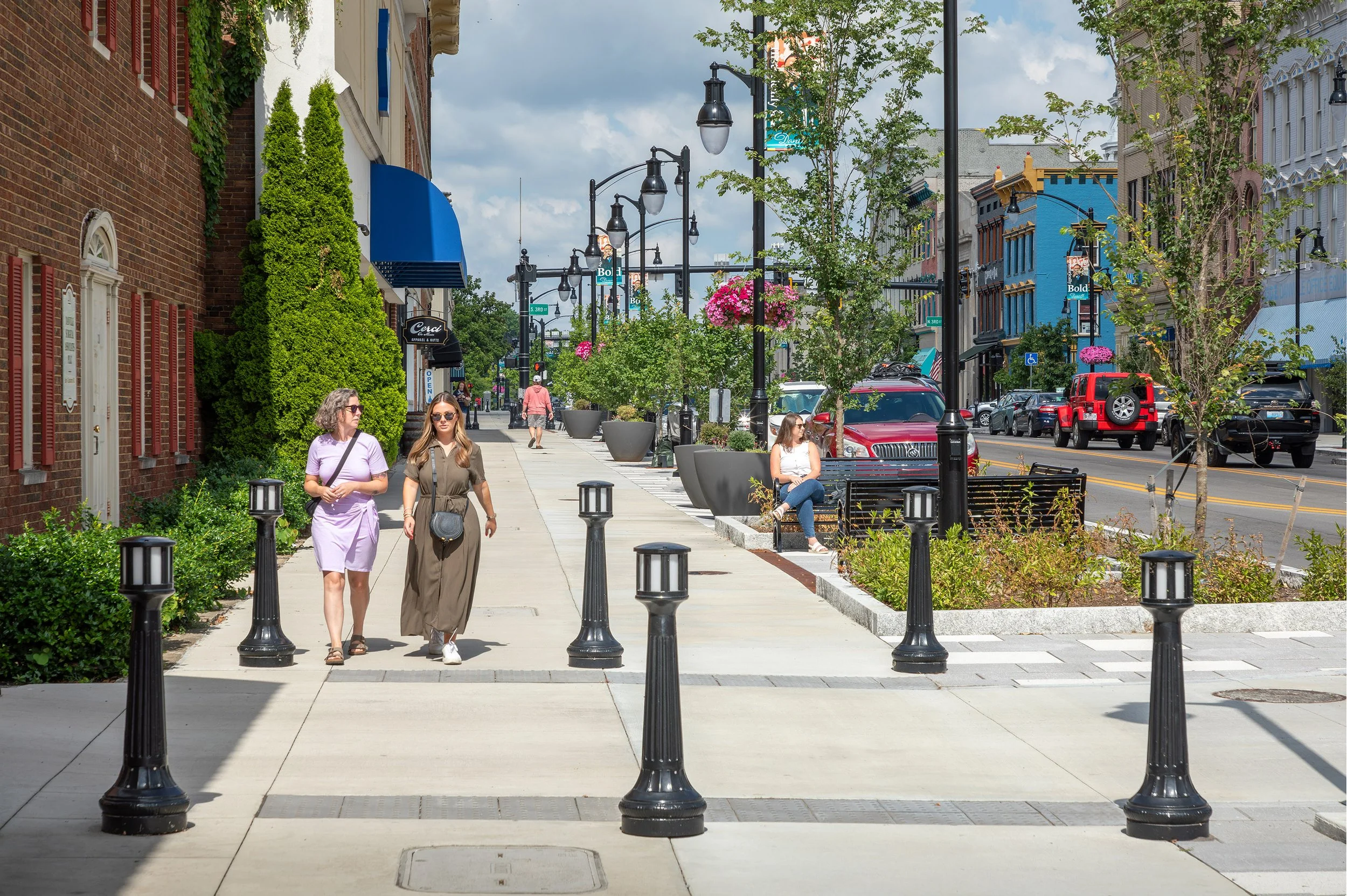 Two women walk along a historic streetscape lined with shops and plants; others sit on benches while cars are parked nearby.