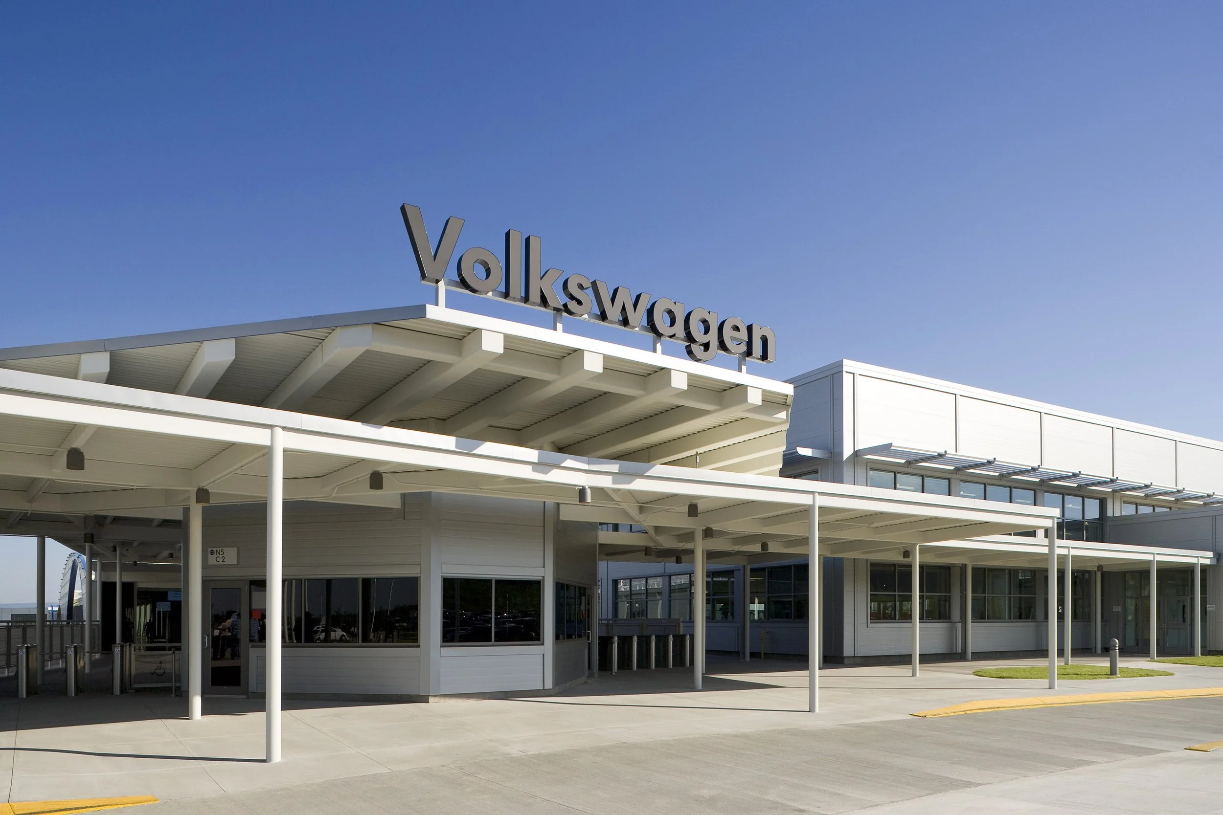 Modern white Volkswagen building with a large rooftop sign, assembly area nearby, under a clear blue sky.