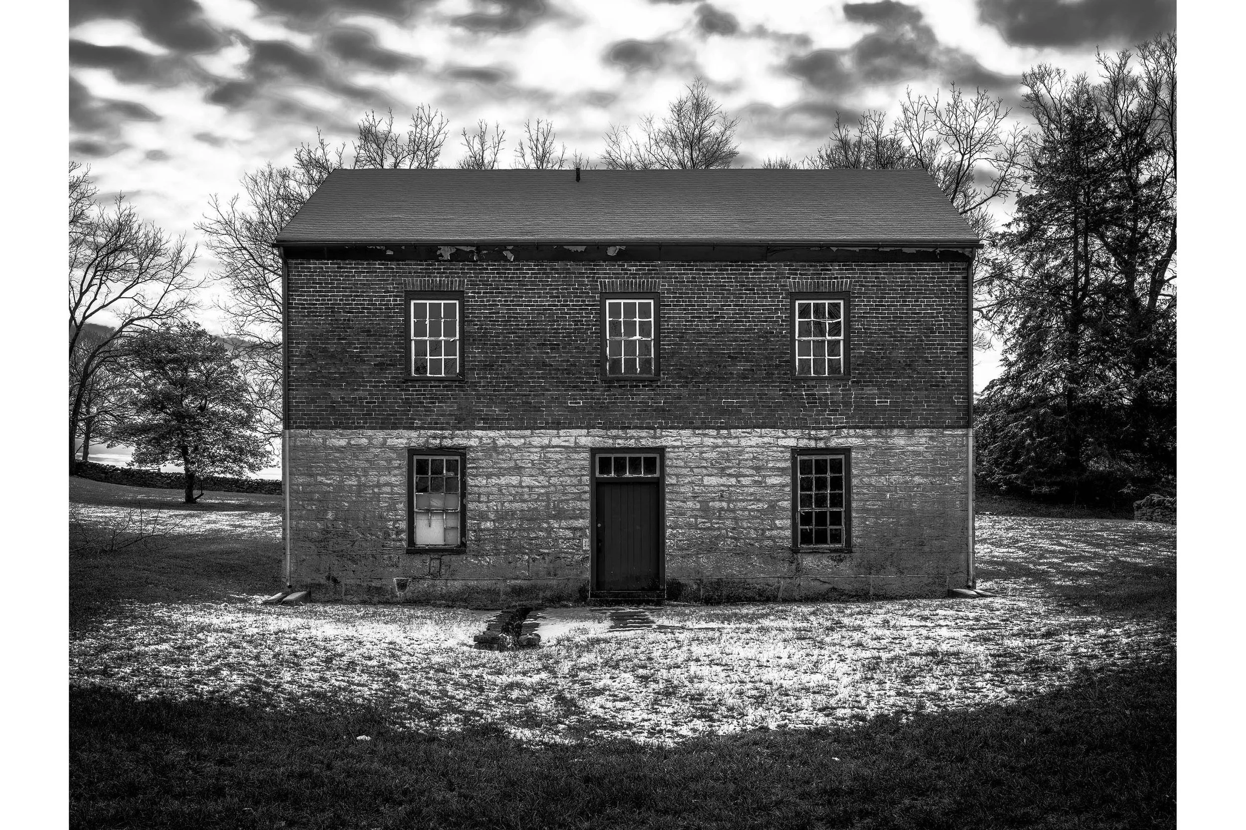 Black and white photo of a Shaker two-story brick and stone house in a winter landscape with leafless trees in the background.