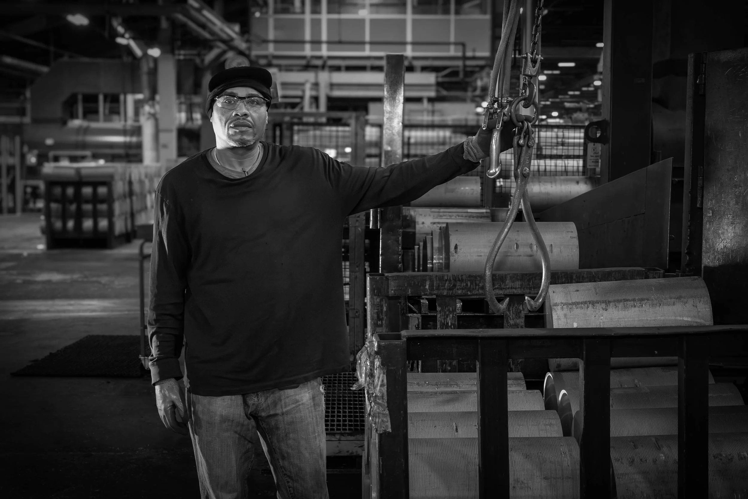 Man standing in a black and white factory, wearing glasses and a cap, with one hand resting on aluminum industrial machinery.