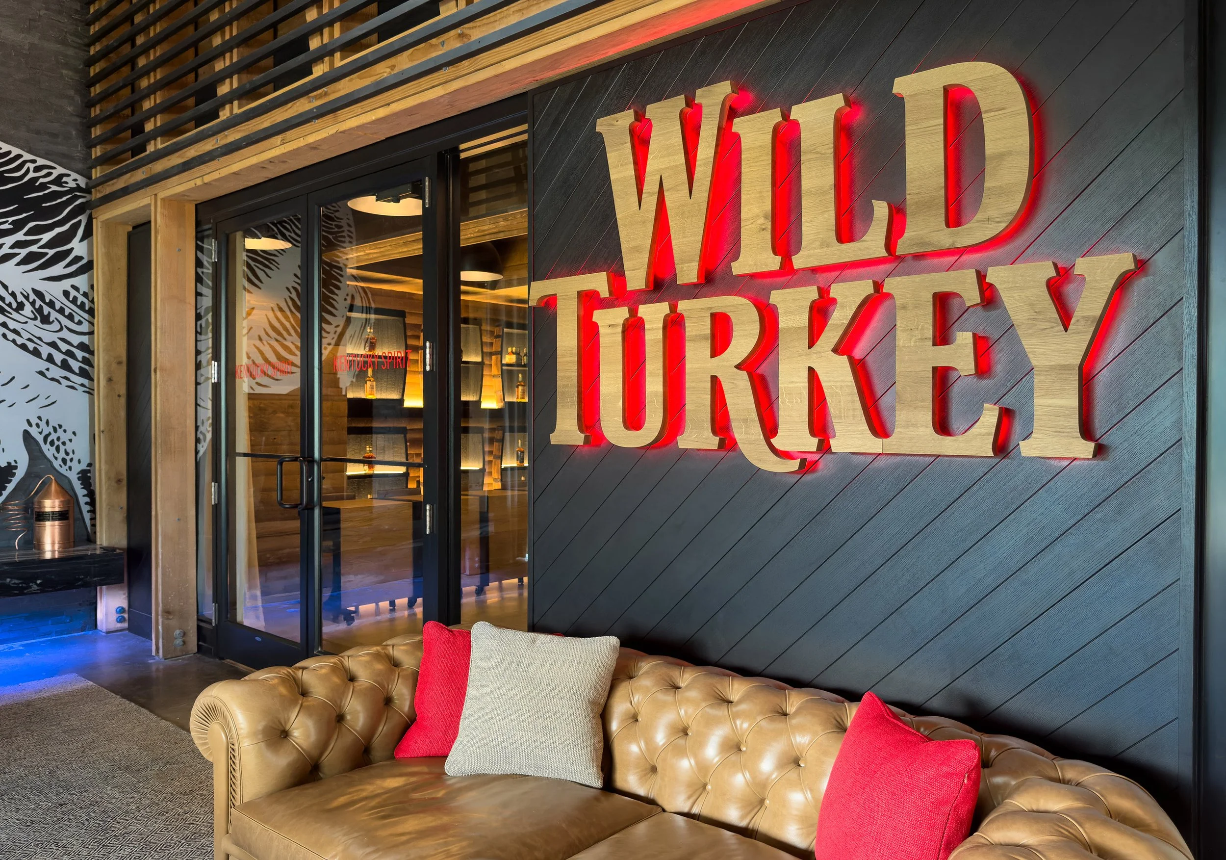 A tan leather couch with red and beige pillows sits in front of a black wall with a large WILD TURKEY sign near glass doors and modern decor.