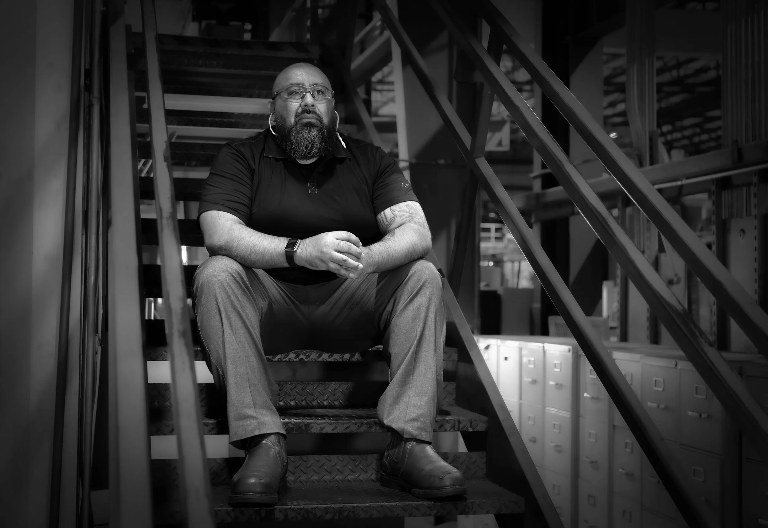 A bearded man sits on aluminum stairs in an industrial setting, looking thoughtful, as if contemplating the manufacturing process. 