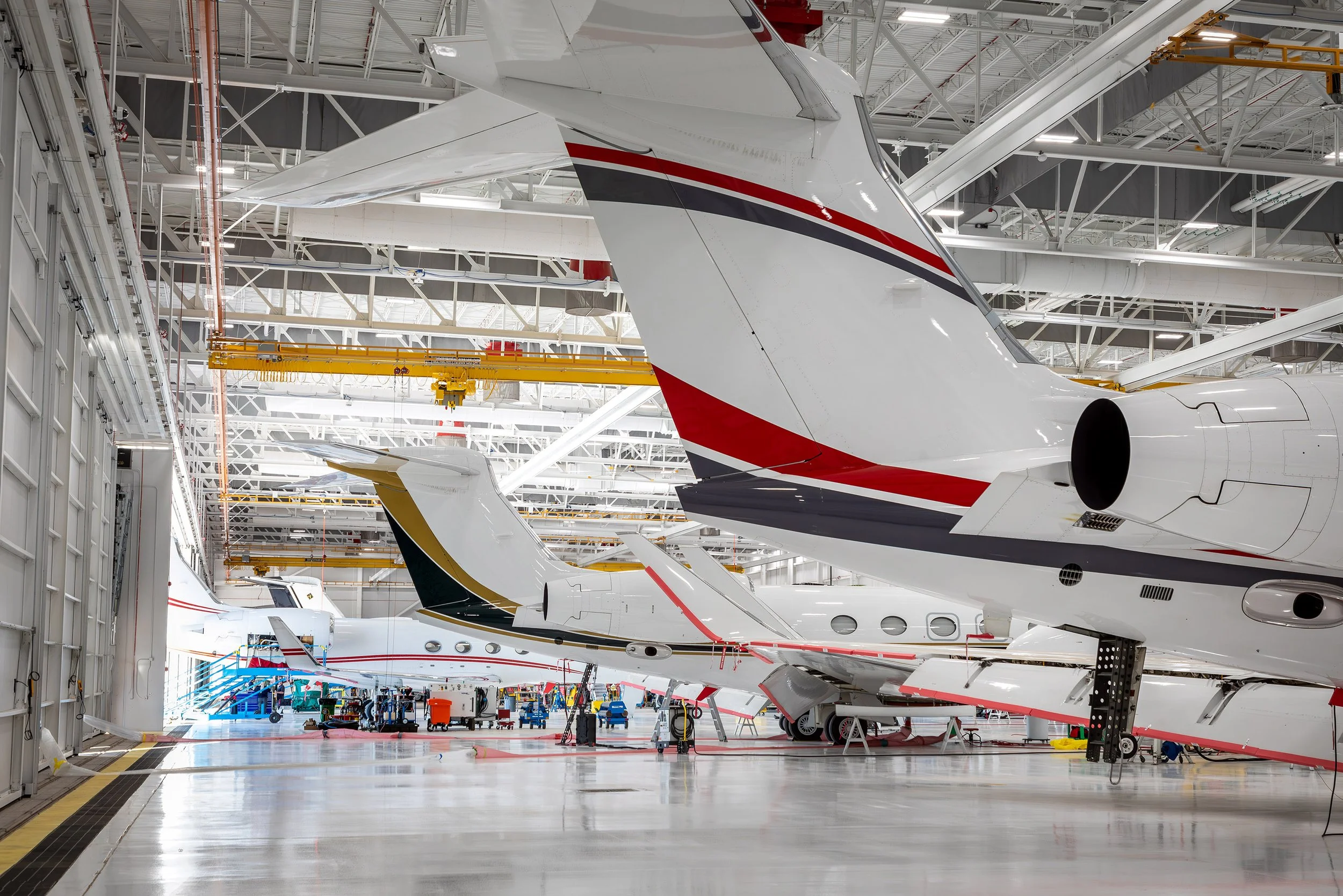 Several private jets are parked inside a spacious, brightly lit aircraft hangar, showcasing impressive architectural features. Workers and equipment are visible around the planes, with the image focusing on the tails and landing gear.