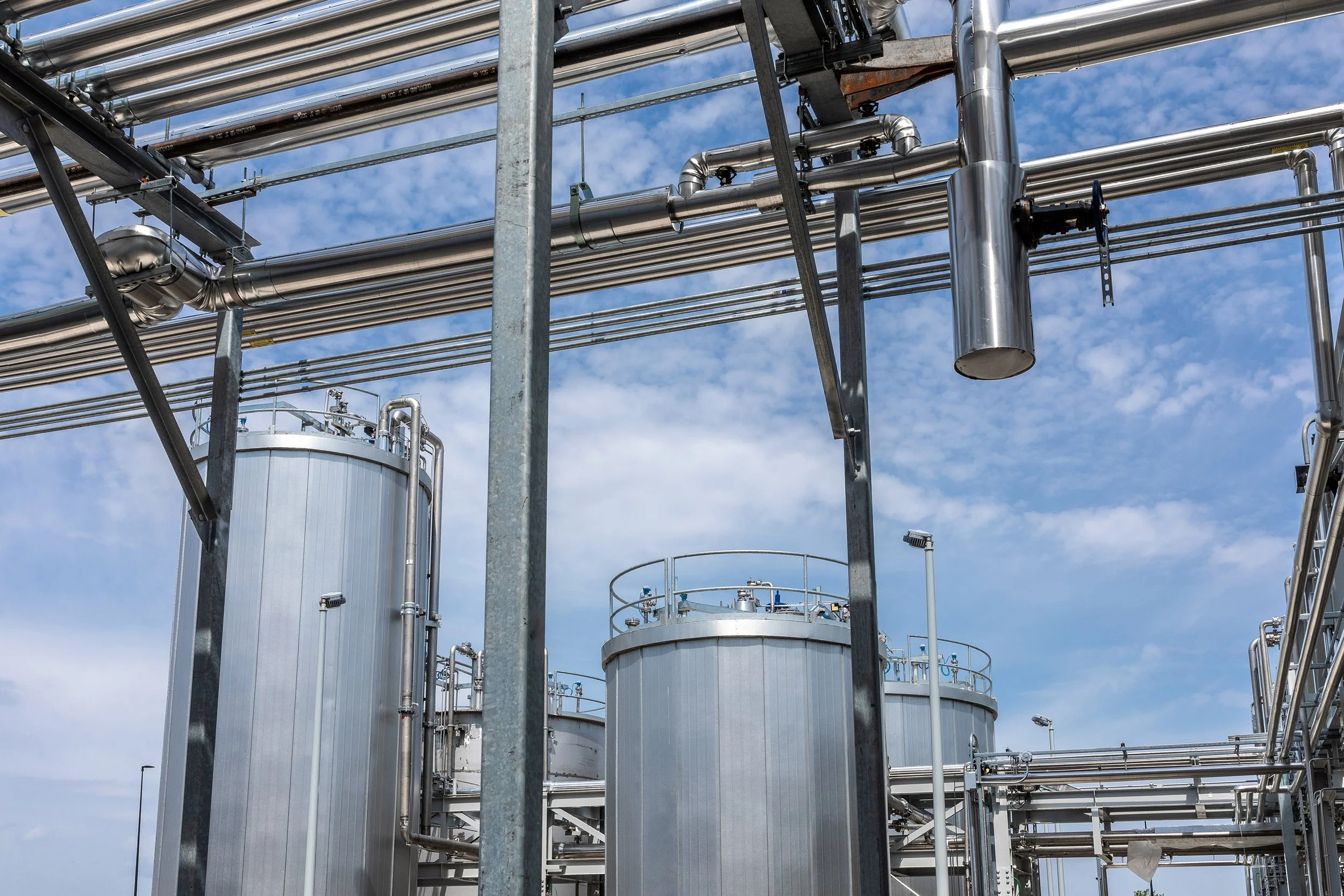 Large, shiny industrial storage tanks and metal pipes are shown against a blue sky with scattered clouds, suggesting an industrial process or manufacturing plant setting.