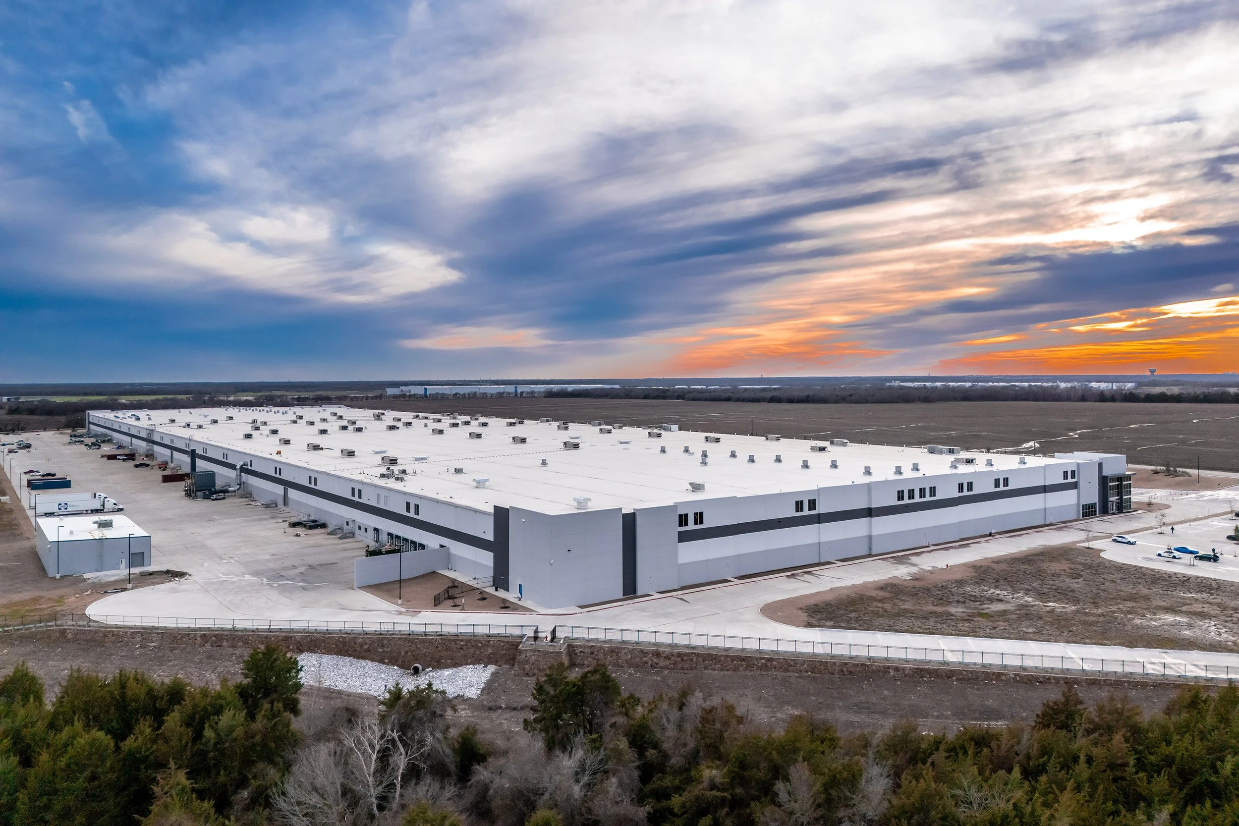 A large, modern industrial warehouse building is surrounded by parking lots and open land under a dramatic, cloudy Texas sunset sky. Sparse trees line the foreground, and open fields extend into the distance.