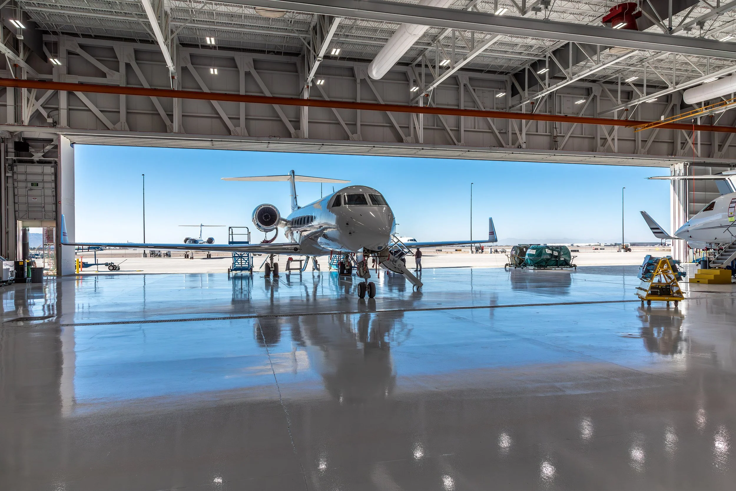 A private jet is parked inside an aircraft hangar in Arizona with its nose facing out toward a sunny runway. Several workers and equipment are positioned near the plane. The shiny, reflective floor adds to the impressive scene—perfect for photography