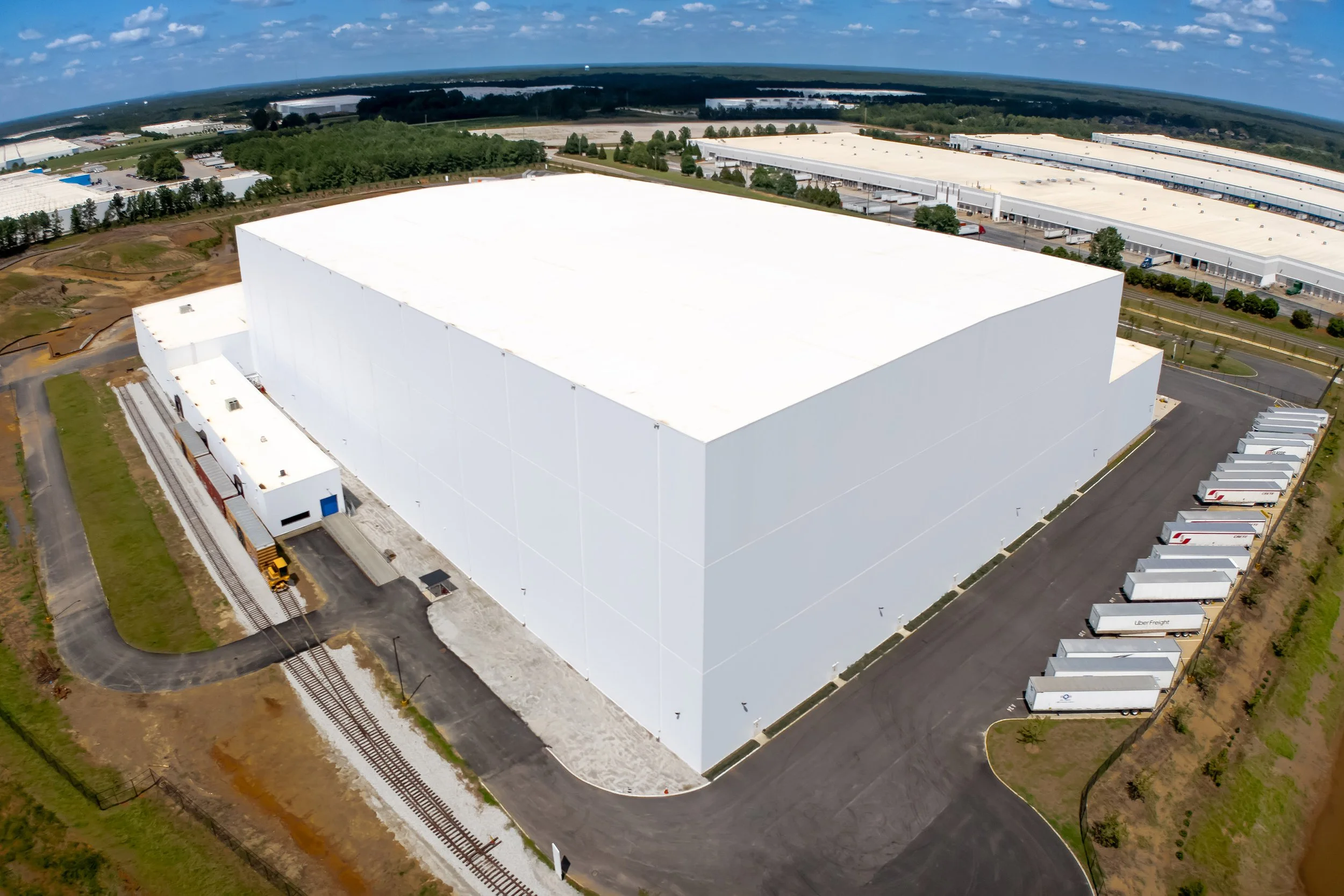 A large white industrial warehouse with loading docks and parked trucks, captured in a detail with landscape and trees in the background.