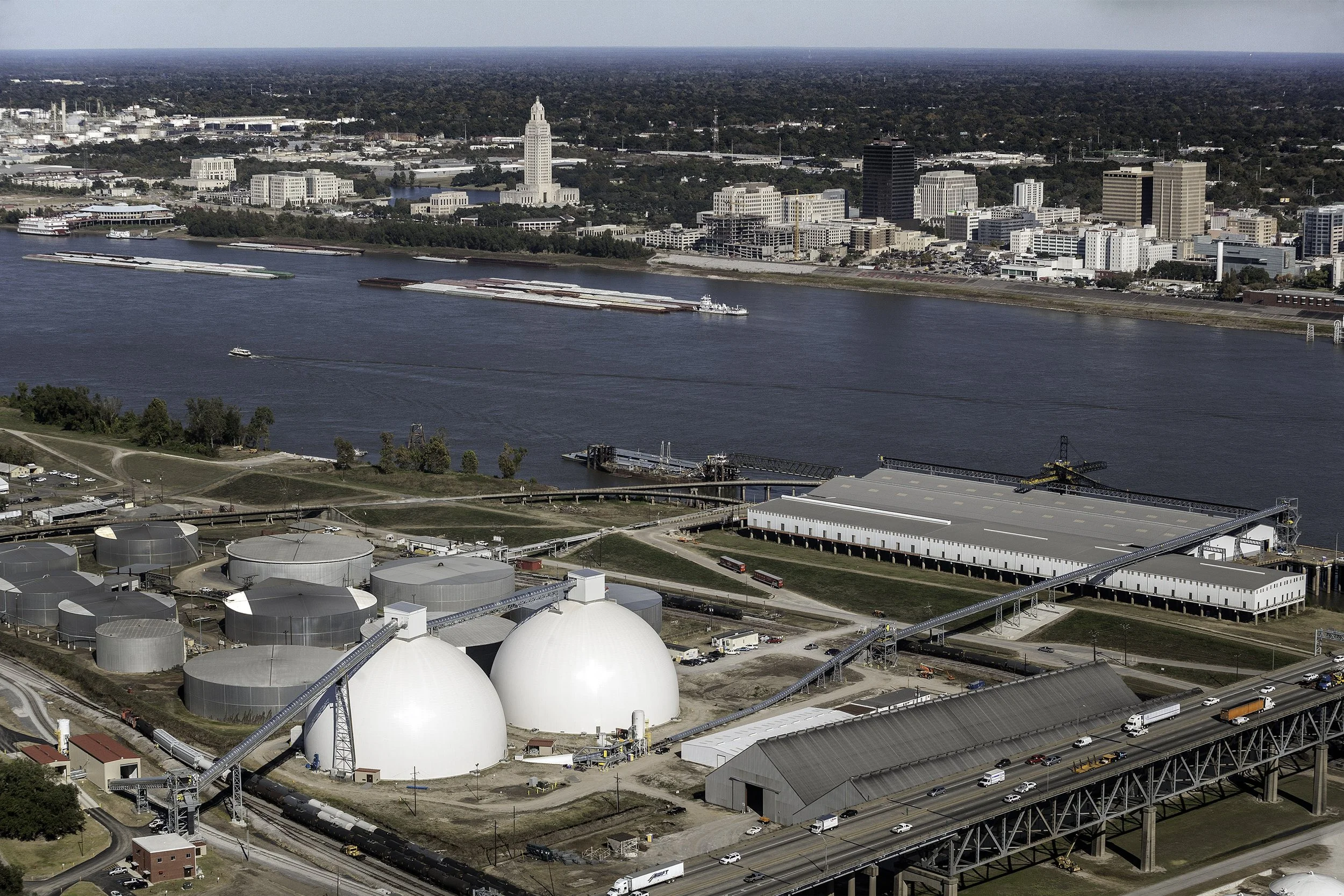 Aerial view of industrial storage domes by a river, with a city skyline and barges in the background.