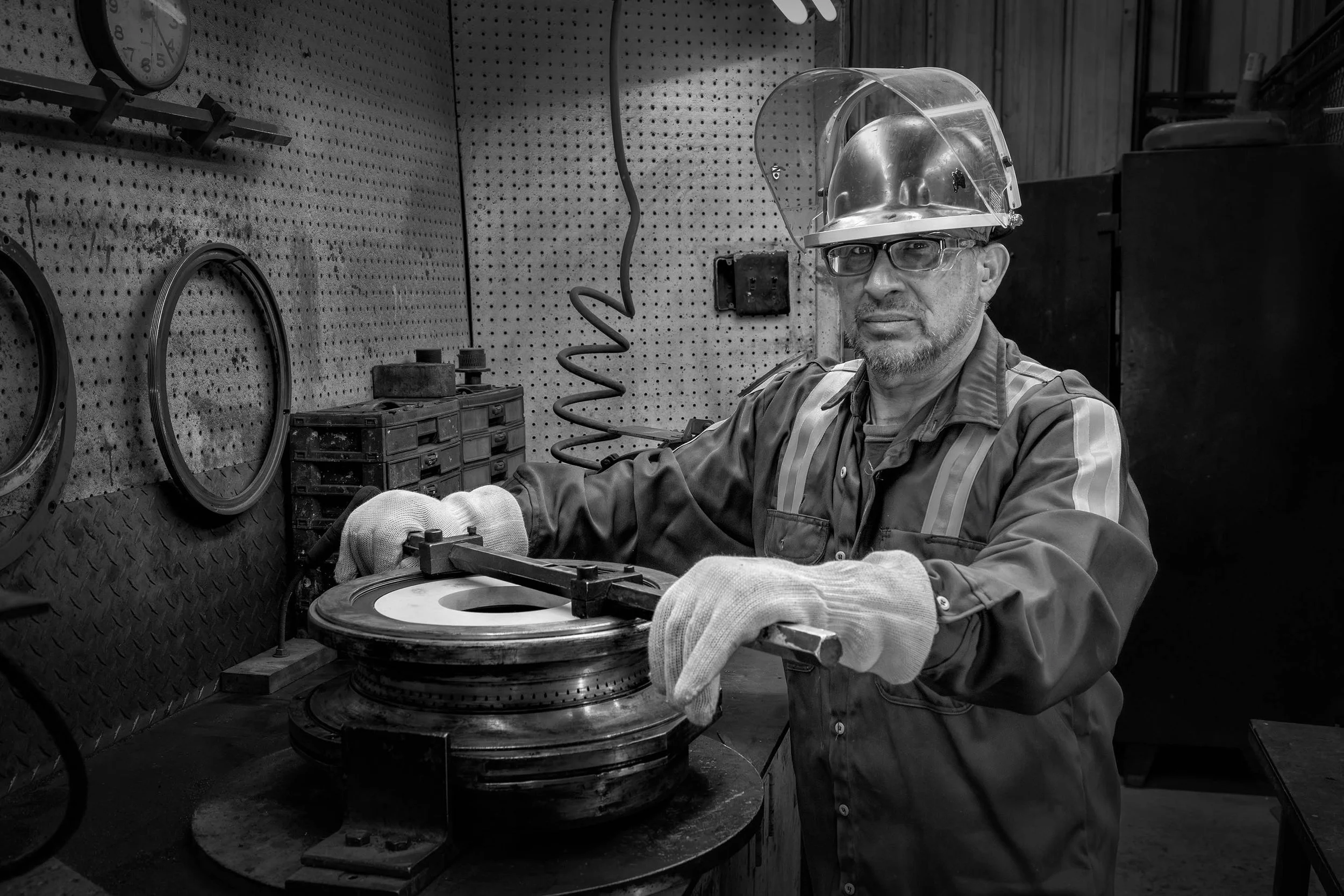 Man in safety gear works with machinery in a black and white industrial workshop, wearing gloves and a face shield.