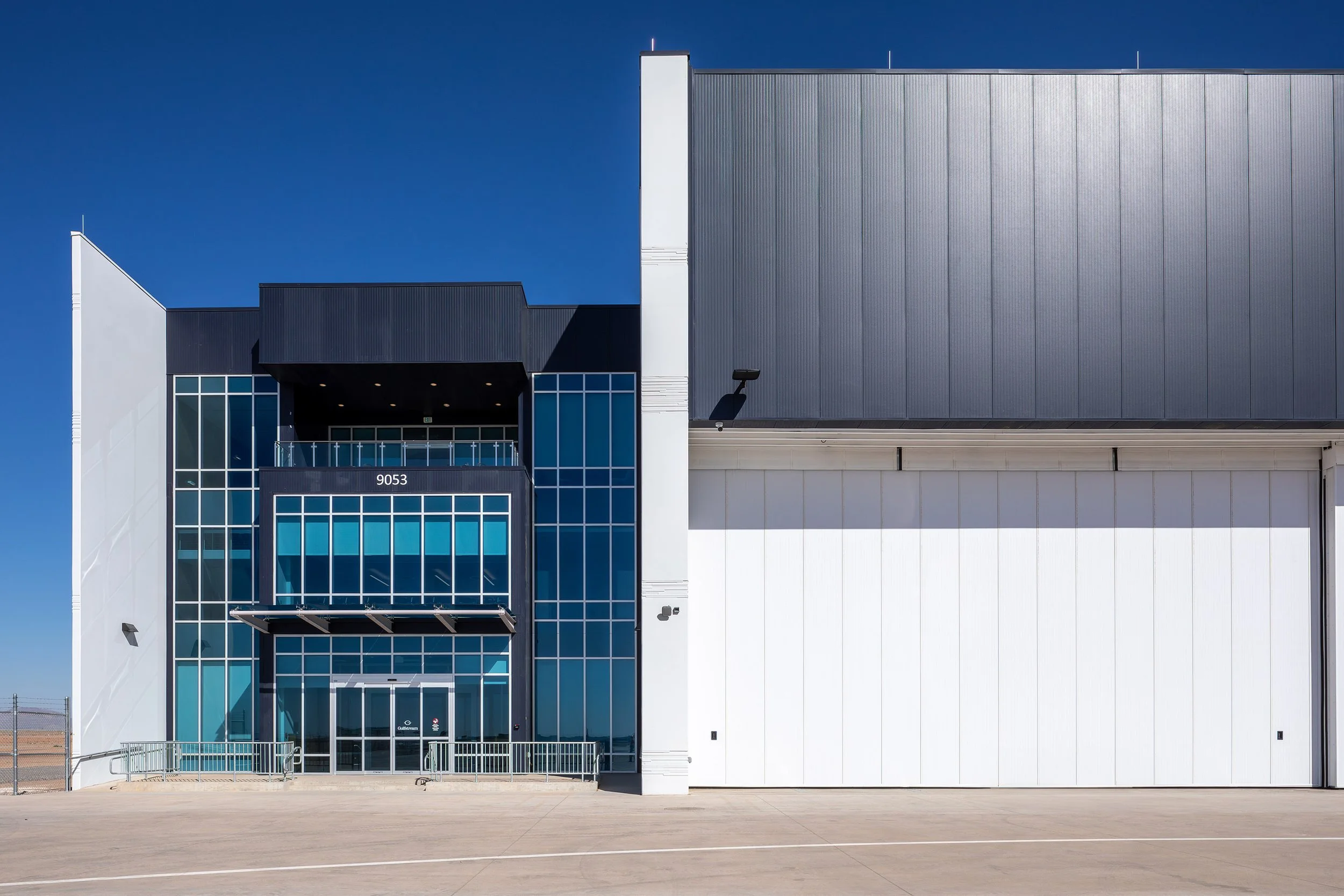 Modern industrial building with a wide white facade, large glass windows, and a central entrance labeled 9053, under a clear Arizona sky. The paved area in front is empty—perfect for photography or aviation-related activities.