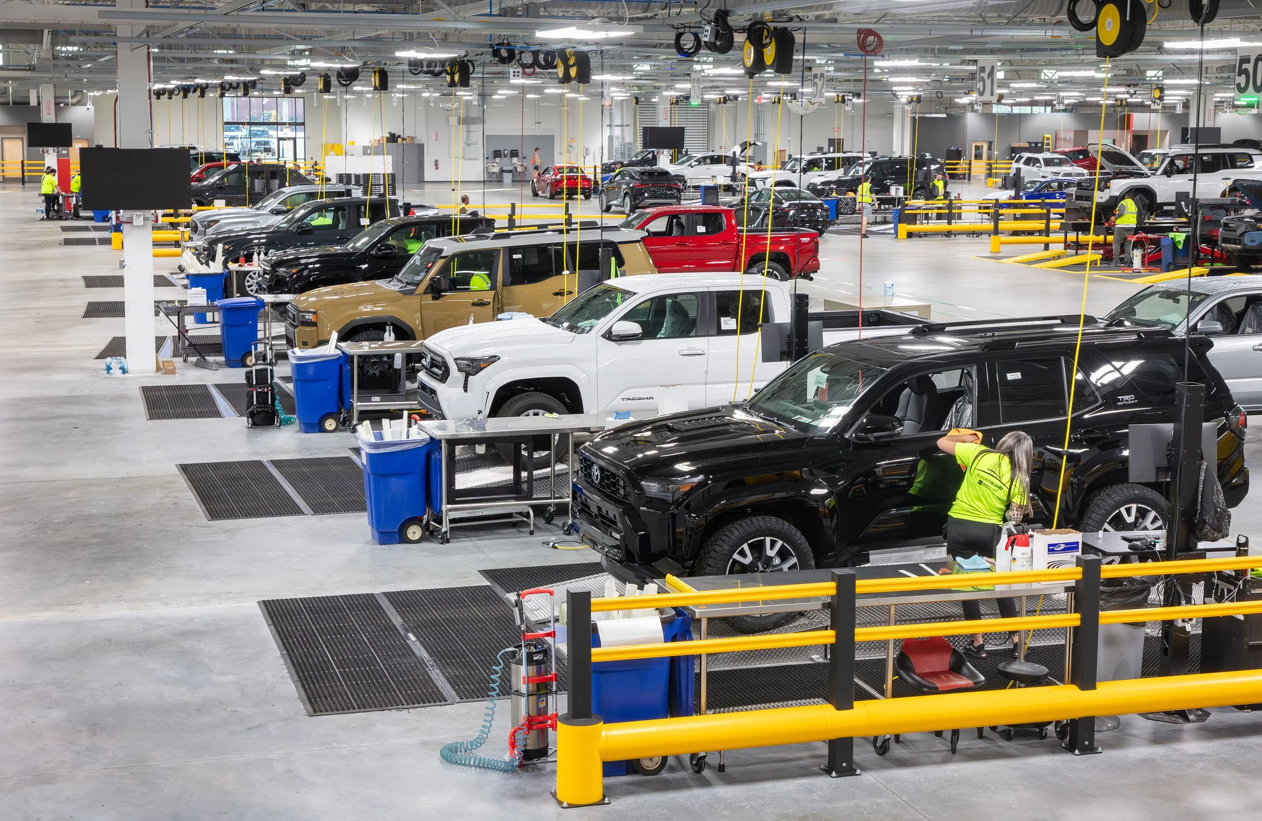 Factory workers inspect SUVs and trucks lined up in a brightly lit, modern automotive assembly plant, with workstations, tools, and supply carts visible throughout the spacious facility.