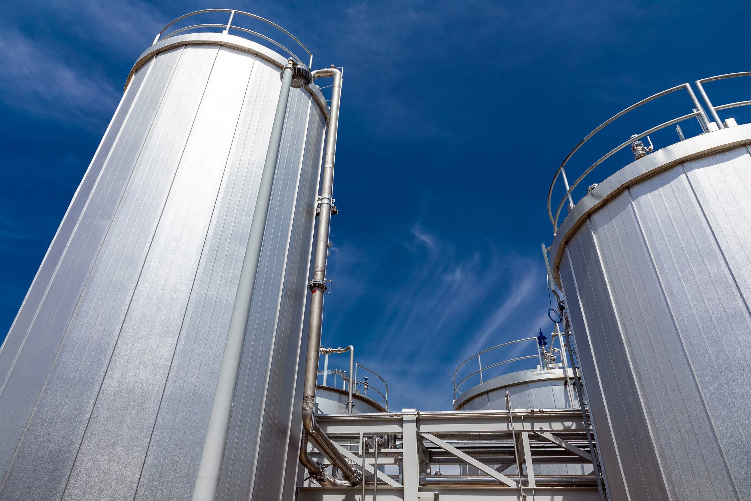 Three large, shiny industrial storage tanks with ladders and railings stand against a clear blue sky with wispy clouds, highlighting the process infrastructure from a low angle.