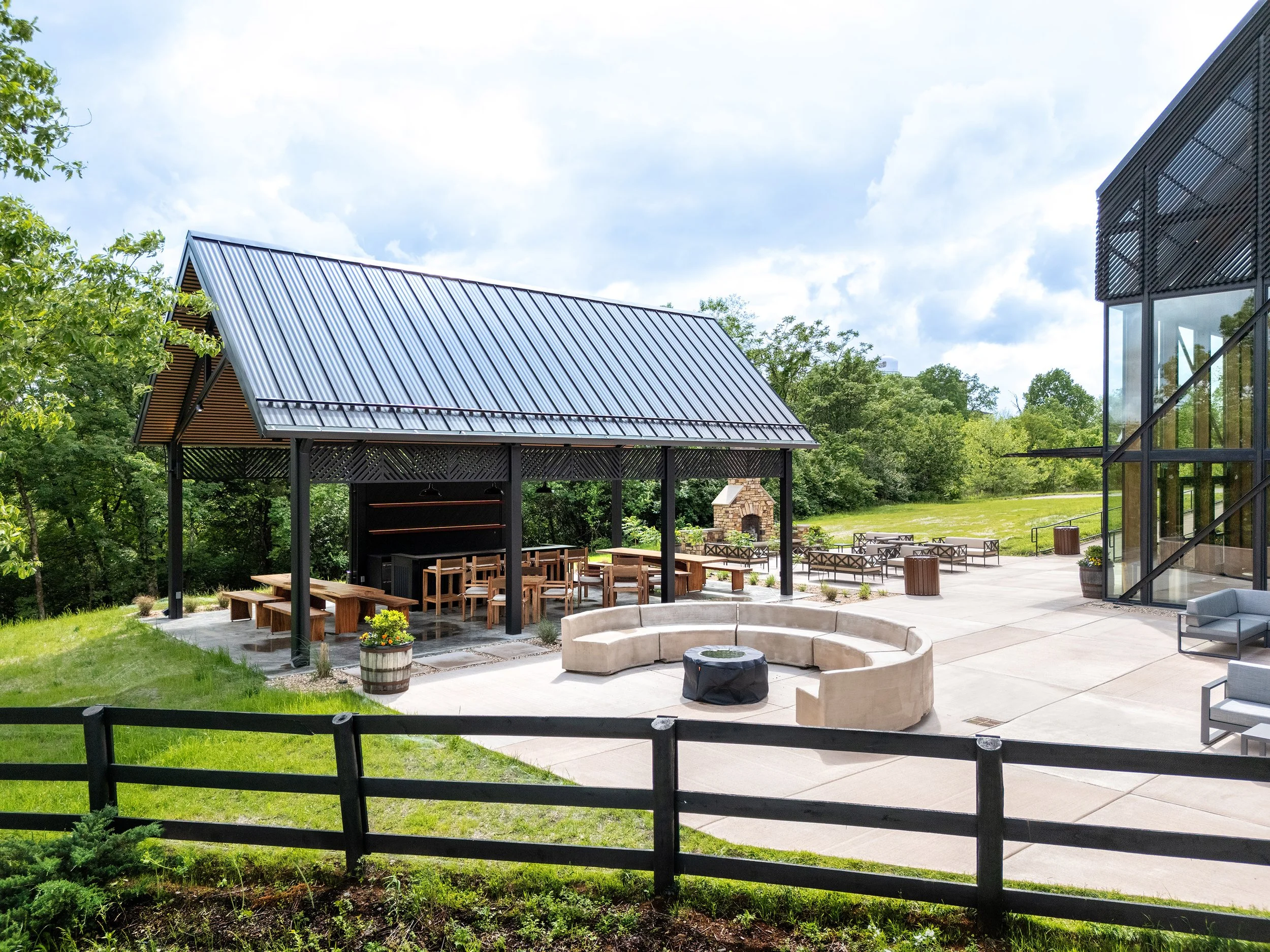 A modern outdoor picnic area with a metal-roofed pavilion, wooden tables, a circular stone fire pit, and glass-walled visitor center, surrounded by green grass and trees under a partly cloudy sky—perfect for photography.