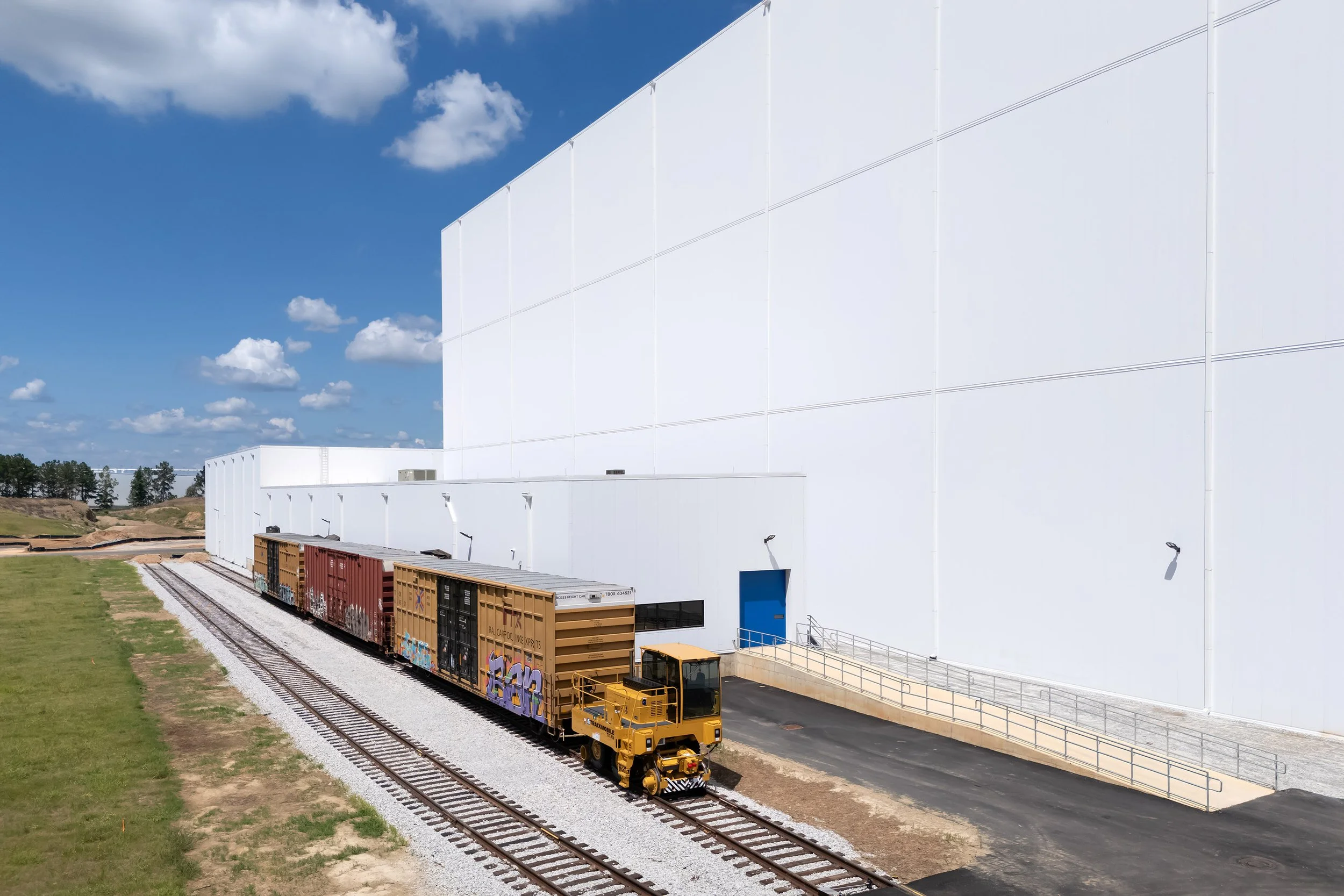 A small yellow train engine pulls freight cars along tracks beside a Georgia industrial refrigeration building under a blue sky.