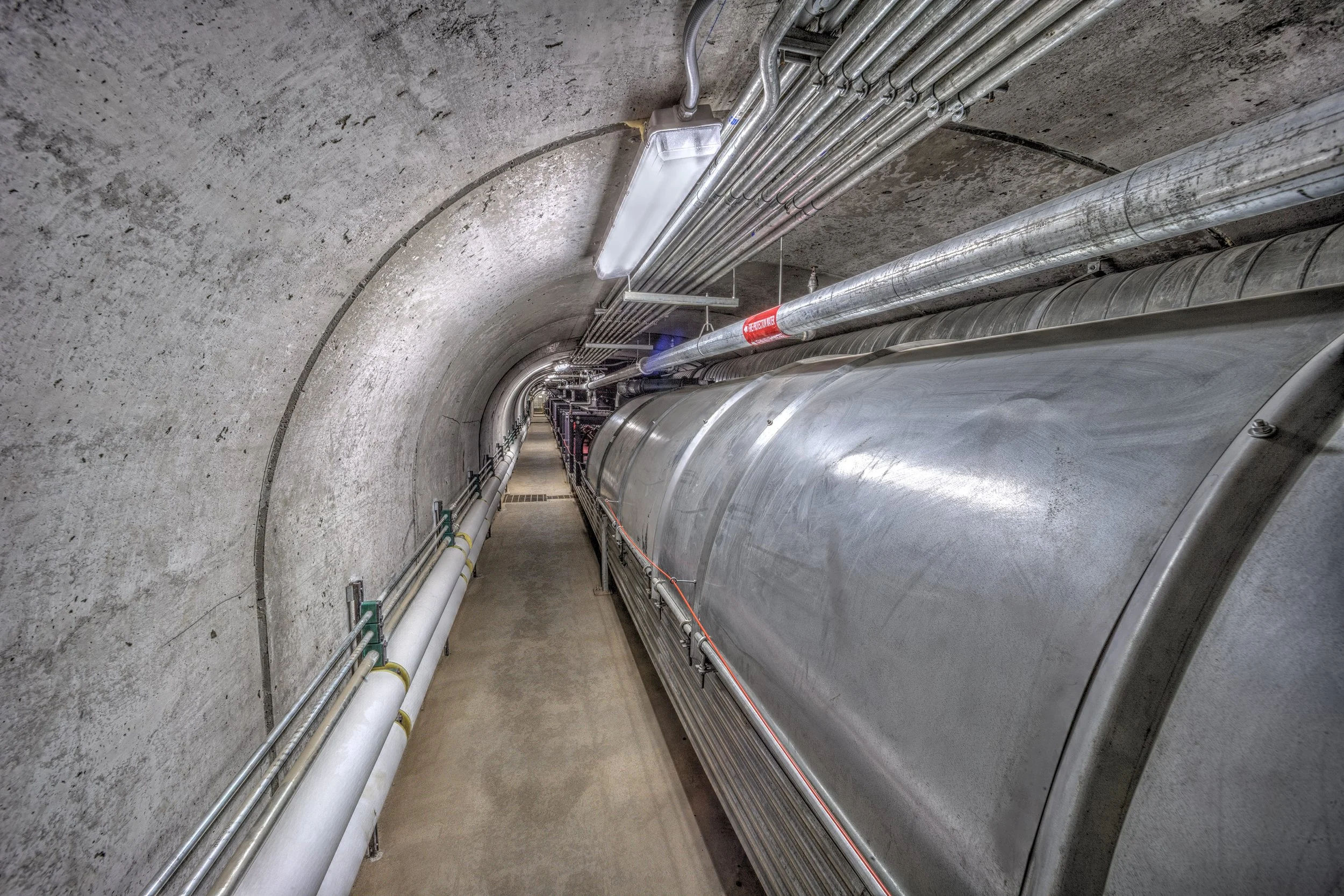 A concrete tunnel with large metal pipes and cables running along the walls and ceiling.