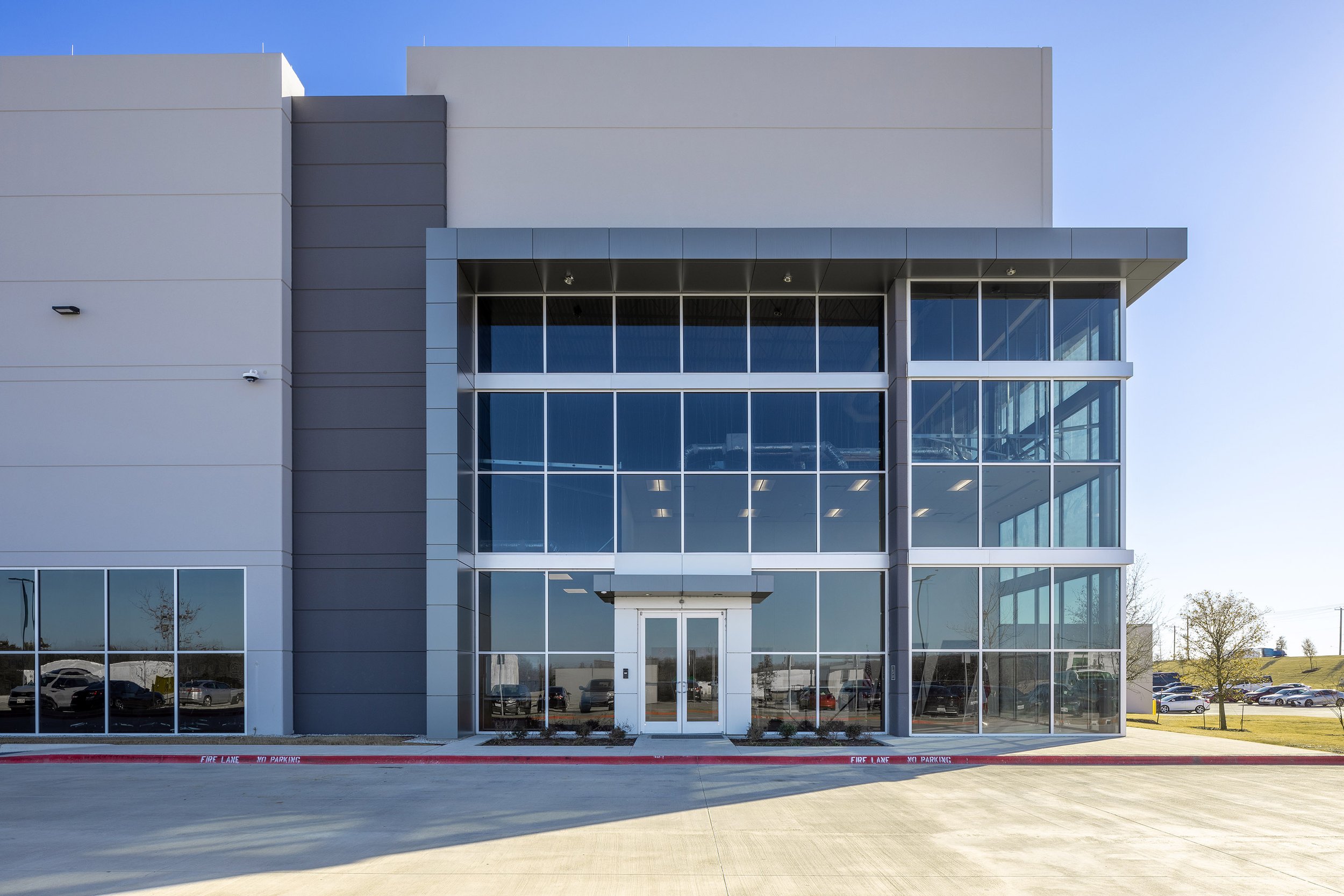 Modern industrial commercial building in Dallas featuring large floor-to-ceiling glass windows and a central glass entrance, surrounded by a concrete driveway and set against a clear blue sky.