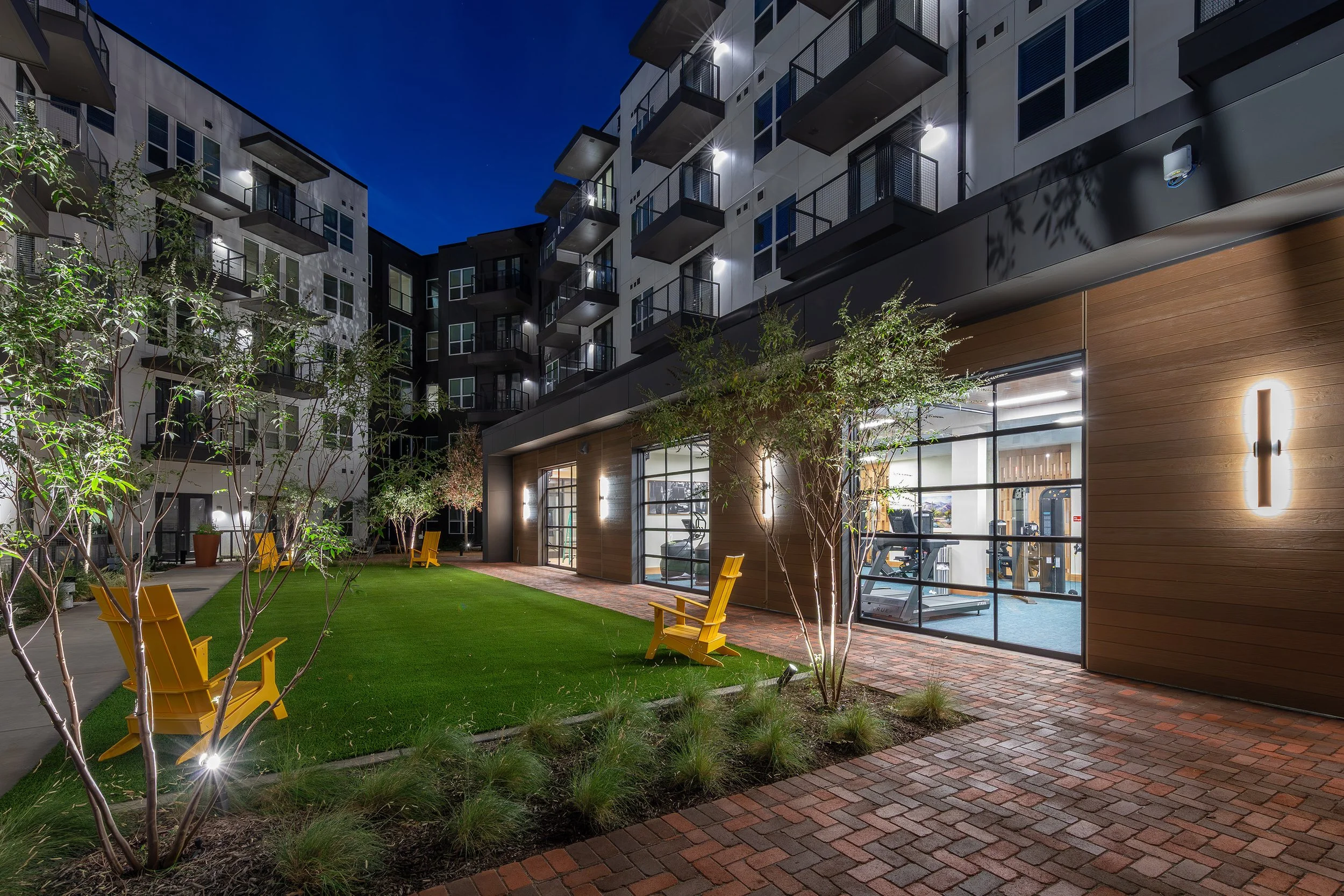 Modern apartment building courtyard at night featuring yellow chairs on a grassy area, brick pathways, young trees, and large windows revealing a well-lit gym inside.
